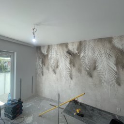 MUR-BETON - Pokój w trakcie dekorowania ścian tapetą z motywem palm, widoczne narzędzia i sprzęt budowlany, surowy beton na podłodze, światło dzienne z balkonu.