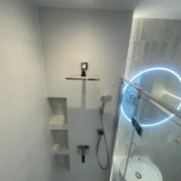 Nowoczesna łazienka po remoncie: szare płytki, wnęki w ścianie, deszczownica, umywalka z podświetlanym lustrem i szklany panel prysznicowy.