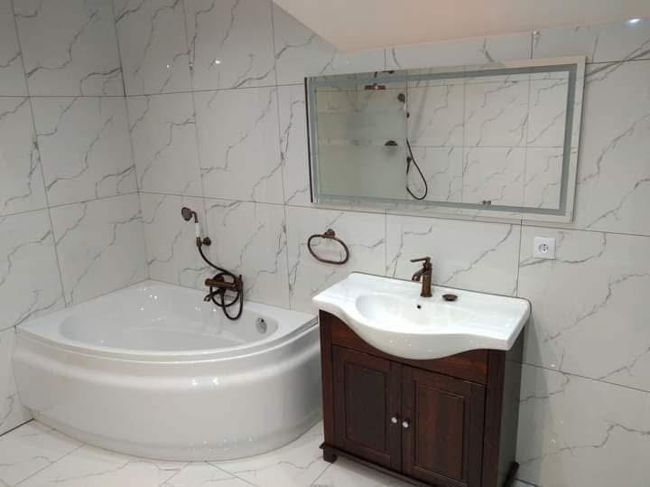 Nowoczesna łazienka z wanną narożną, umywalką na szafce z ciemnego drewna i dużym lustrem z podświetleniem. Białe płytki imitujące marmur na ścianach i podłodze.