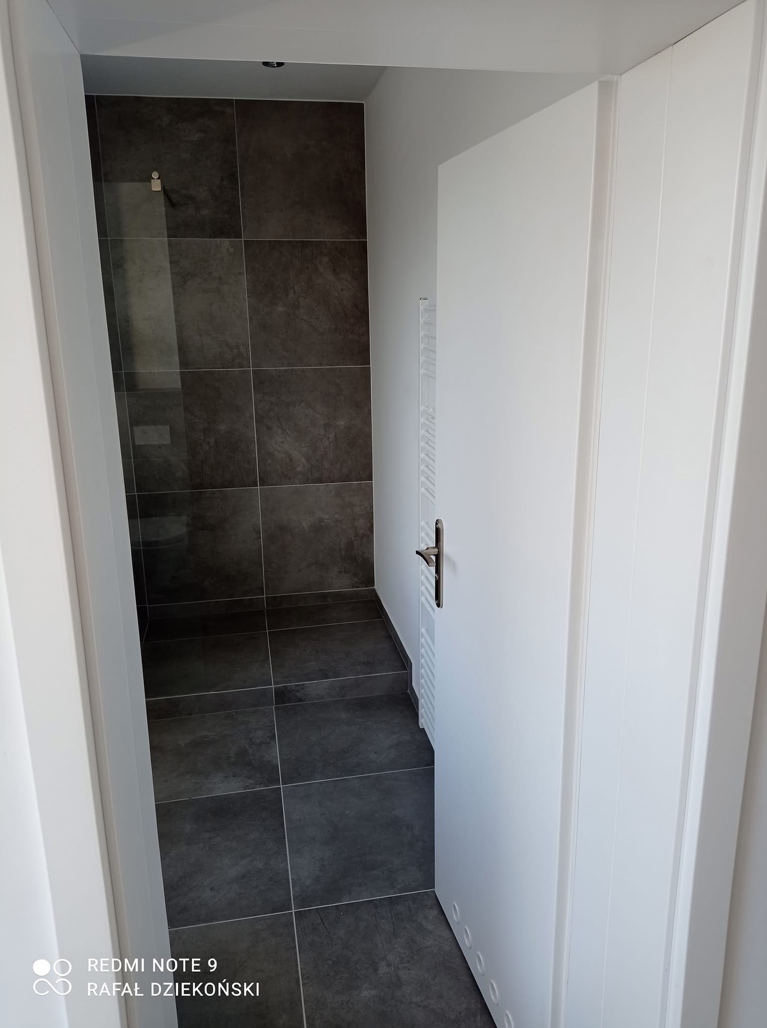 Otwarta biała stolarka drzwiowa, za nią wykończona łazienka z ciemną glazurą i grzejnikiem drabinkowym. Widok na wnętrze przez otwarte drzwi.