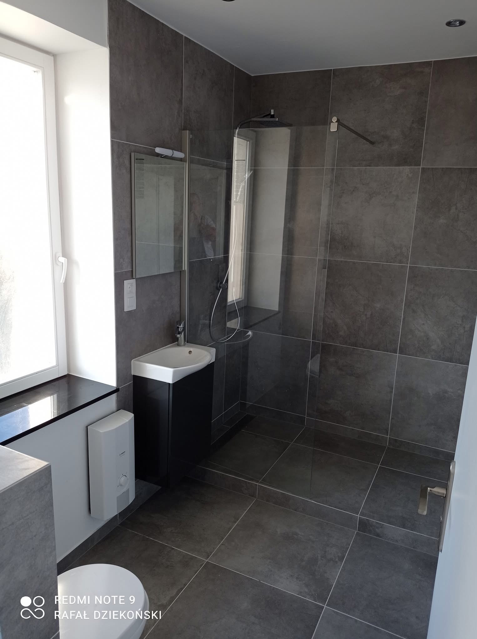 Nowoczesna łazienka w szarościach z kabiną prysznicową typu walk-in, umywalką z szafką i dużym oknem. Minimalistyczny design z płytkami imitującymi kamień.