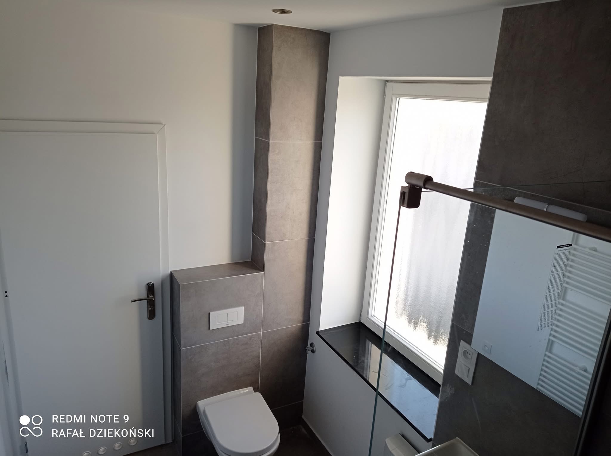 Nowoczesna łazienka po remoncie: szara glazura imitująca kamień, biała armatura i drzwi, okno z czarnym parapetem, minimalistyczne oświetlenie punktowe.