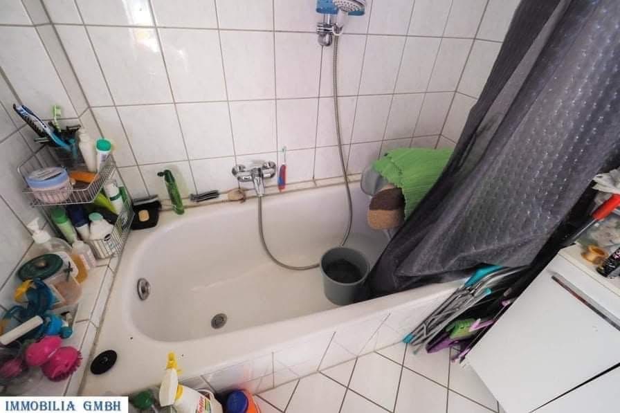 Biała wanna z chromowaną baterią i prysznicem w łazience z białymi płytkami. Widoczne akcesoria łazienkowe i wiadro. Ciemna zasłona prysznicowa w rogu.