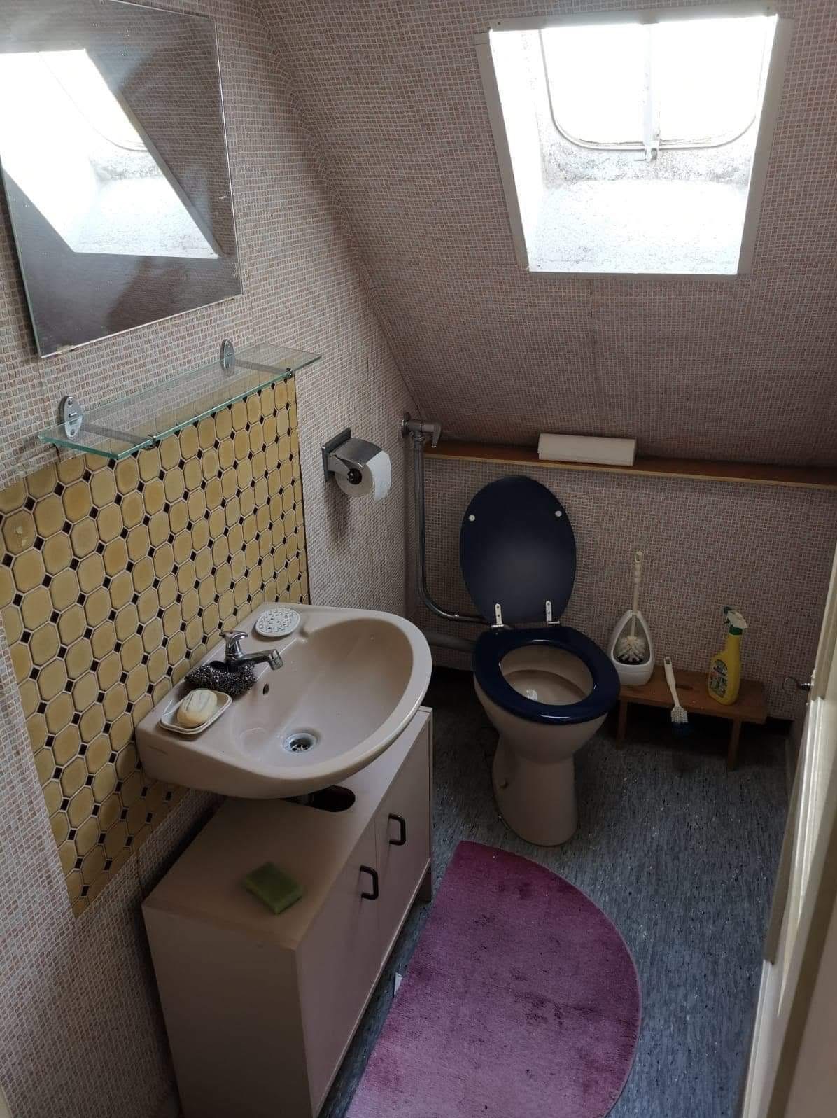 Łazienka na poddaszu z umywalką na szafce, toaletą z ciemną deską i żółtymi płytkami. Widoczne okno dachowe i lustro. Wnętrze o specyficznej estetyce.