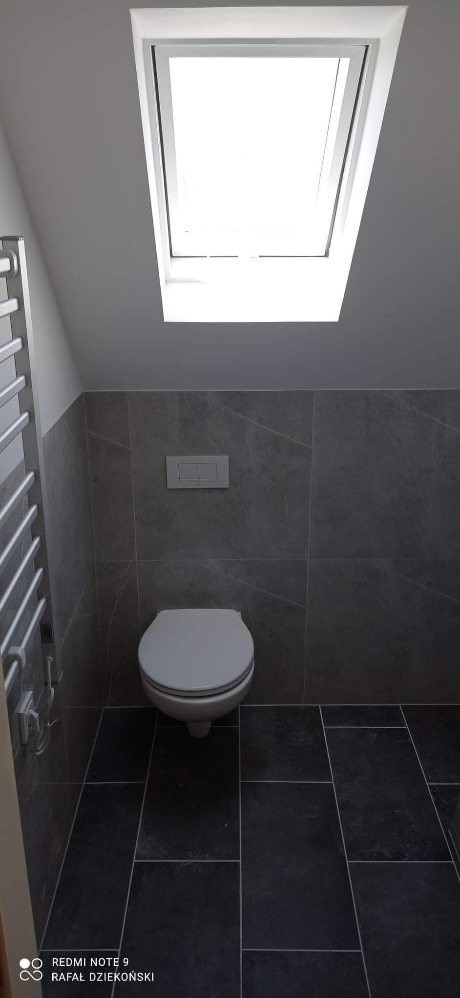 Nowoczesna łazienka na poddaszu z oknem dachowym. Szare płytki imitujące marmur na ścianach i podłodze. Wisząca toaleta z podtynkowym systemem spłukiwania.