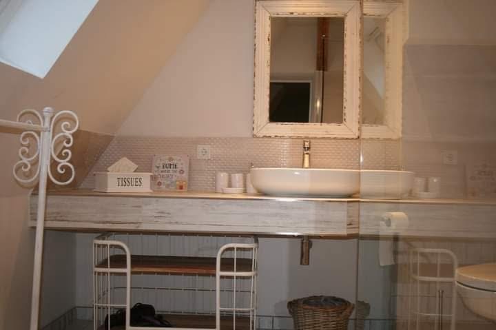 Biała umywalka w stylu vintage z lustrem w ramie, na tle beżowych płytek. Wyposażenie łazienki w jasnych barwach, dekoracyjny regał pod umywalką.