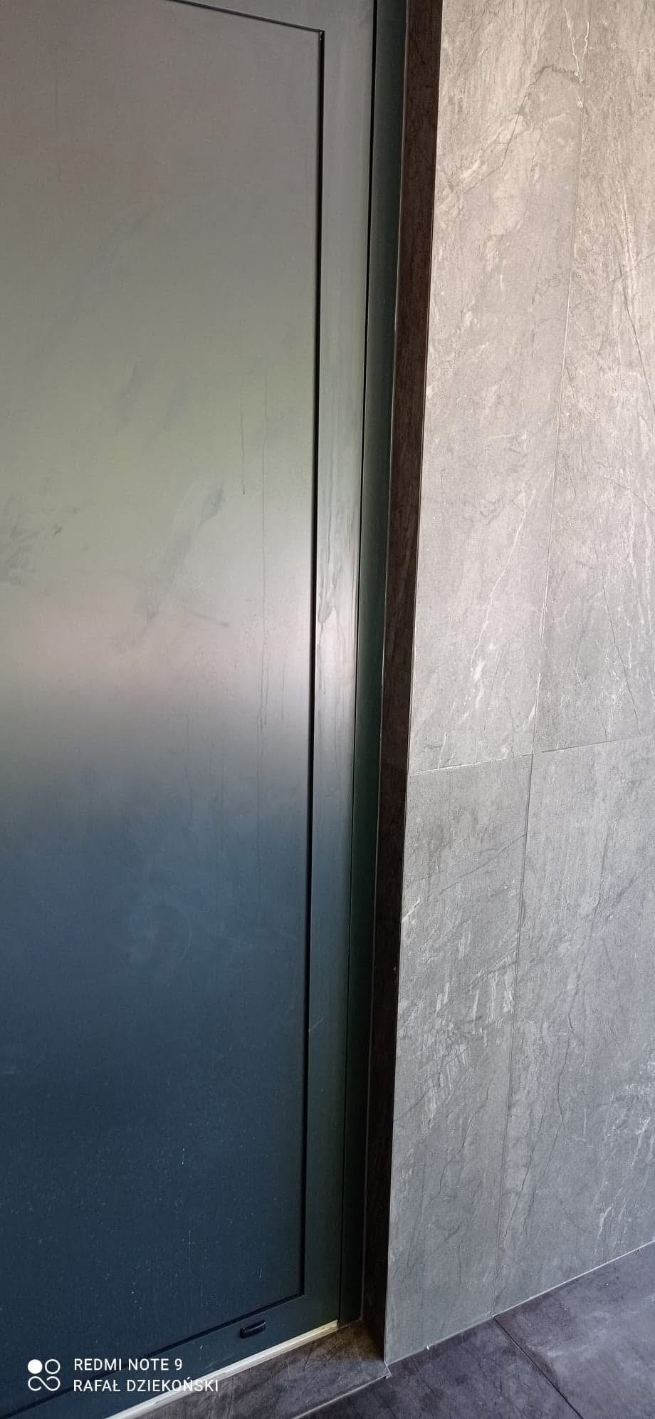 Zamknięte, grafitowe drzwi przesuwne z ciemną ramą, przylegające do ściany wyłożonej szarymi, marmurkowymi płytkami. Minimalistyczny design.