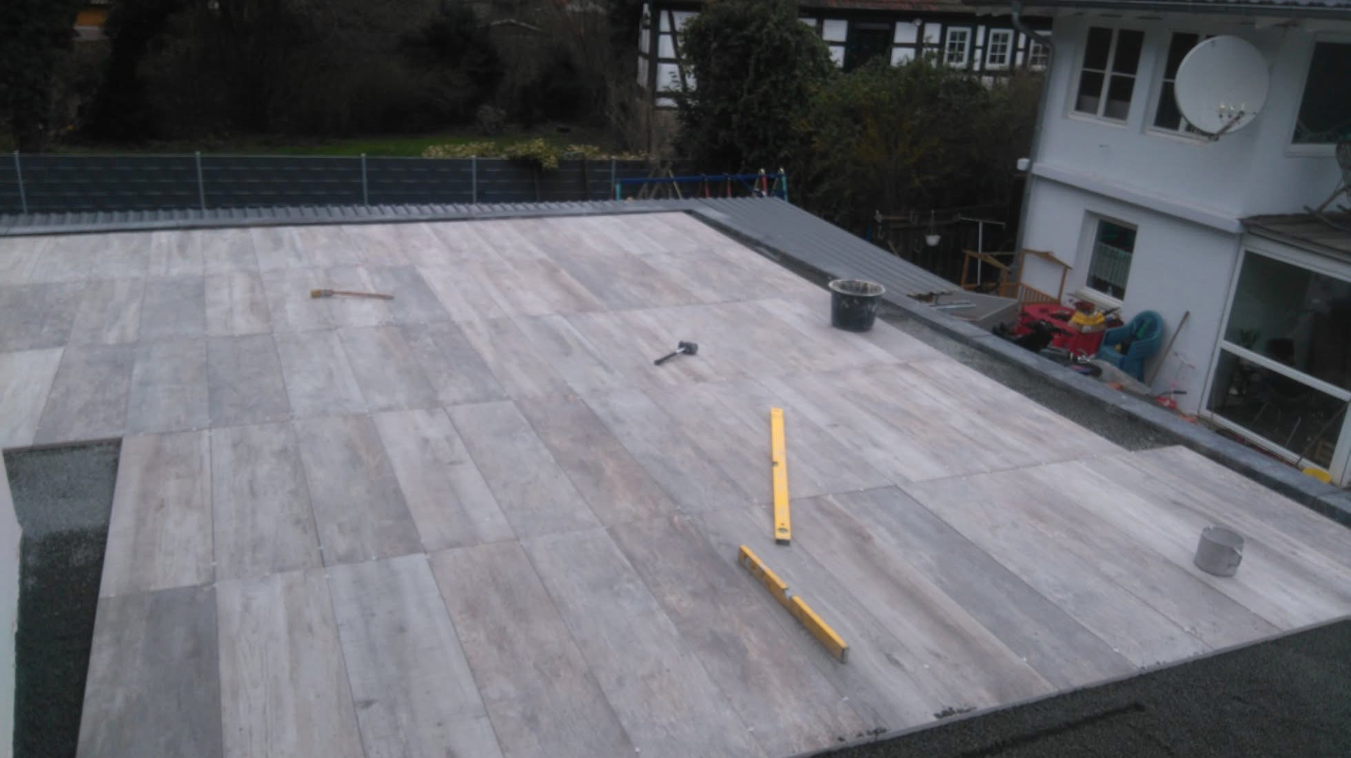 Układanie jasnoszarych płytek na dachu tarasu. Widoczne narzędzia: poziomica, młotek gumowy i wiadro. W tle dom z białymi ścianami i zielonymi drzewami.