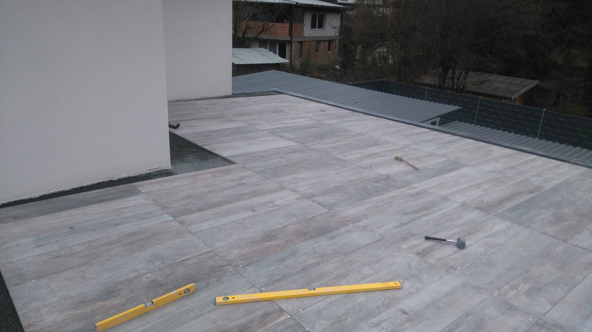 Ułożona szara podłoga z płytek imitujących drewno na tarasie. Widoczne poziomice i młotek. W tle budynki i drzewa.