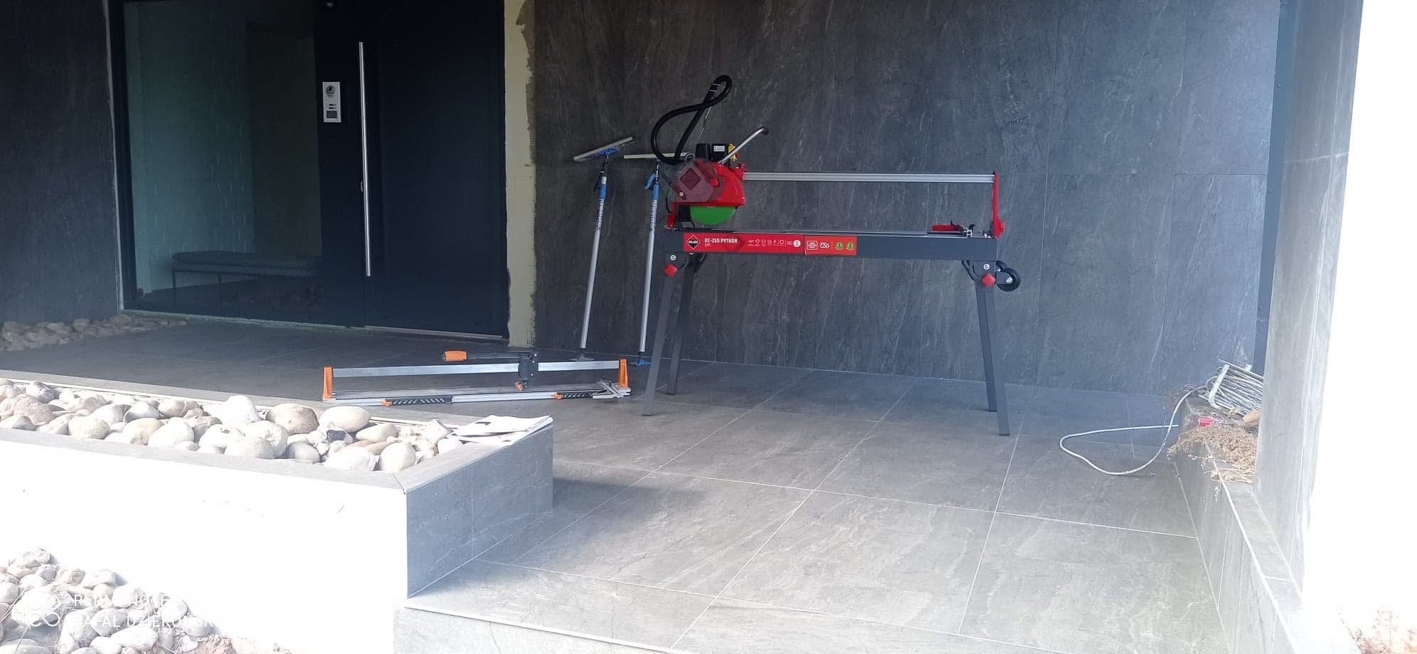 Profesjonalna przecinarka do płytek RUBI DC-250 Python na tle szarej ściany i płytek podłogowych, obok drzwi wejściowych. Widoczne narzędzia glazurnicze.