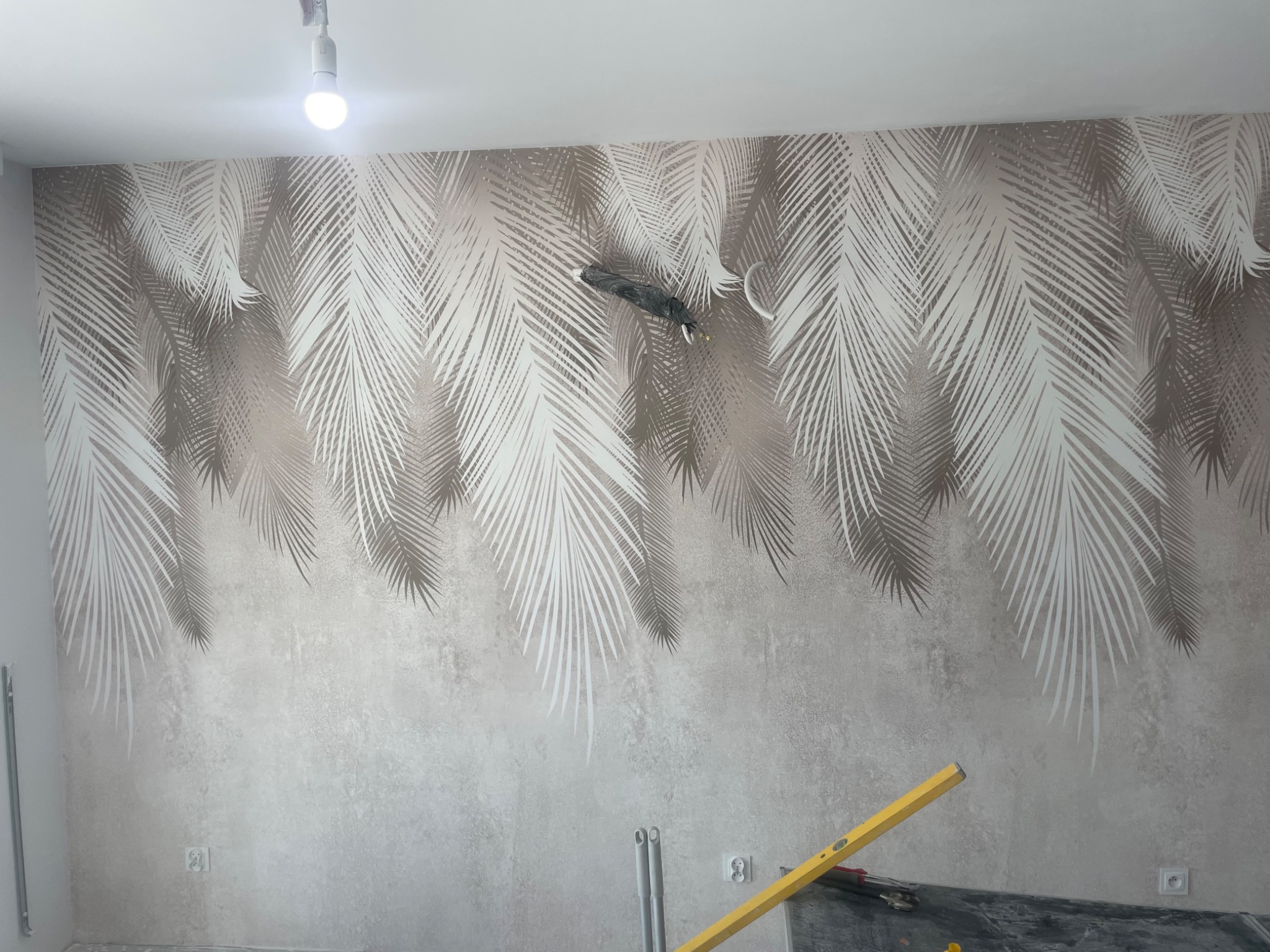 Ściana z tapetą w motywie palmowych liści, na dole widoczna poziomica, sugerująca prace wykończeniowe. Oświetlenie sufitowe podkreśla wzór tapety.