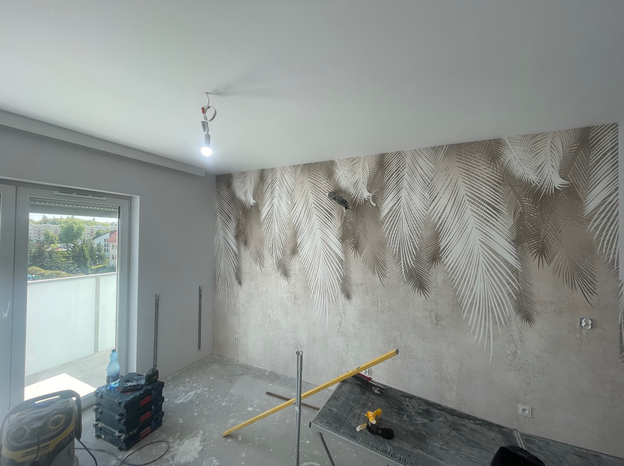 Pokój w trakcie dekorowania ścian tapetą z motywem palm, widoczne narzędzia i sprzęt budowlany, surowy beton na podłodze, światło dzienne z balkonu.