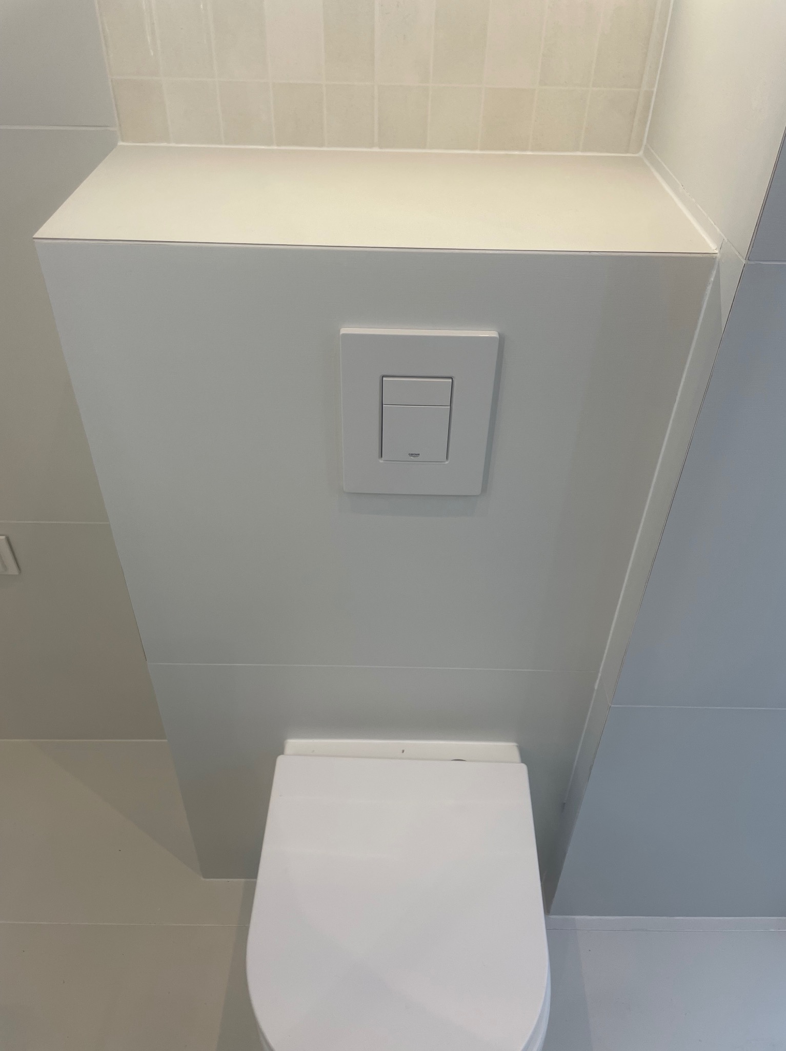 Nowoczesna, podwieszana toaleta z białym przyciskiem Grohe. Jasne płytki na ścianach i podłodze. Minimalistyczny design łazienki.