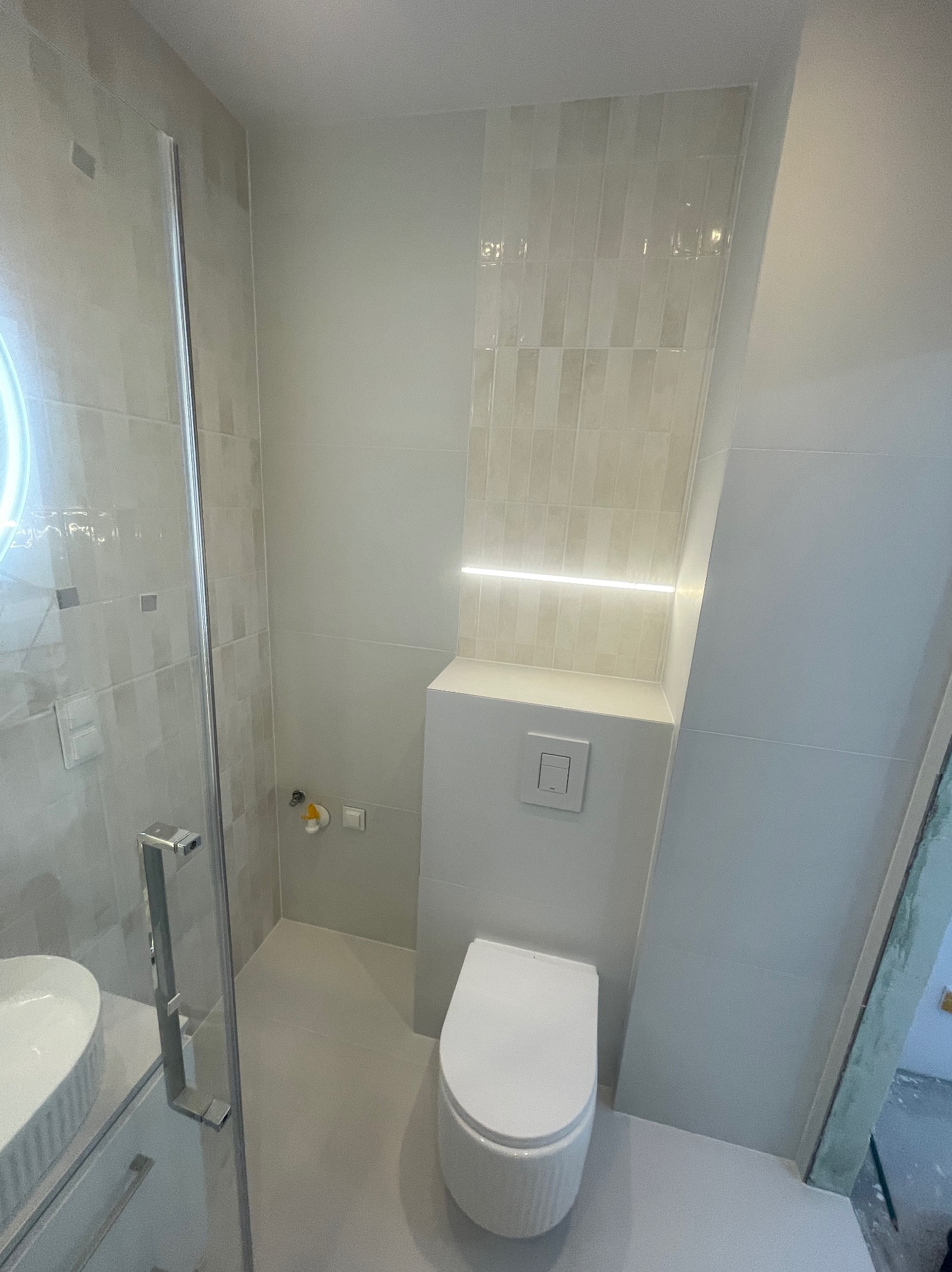 Jasna łazienka po remoncie: kabina prysznicowa ze szklanymi drzwiami, podwieszana toaleta, płytki w odcieniach beżu, oświetlenie LED. Minimalistyczny design.