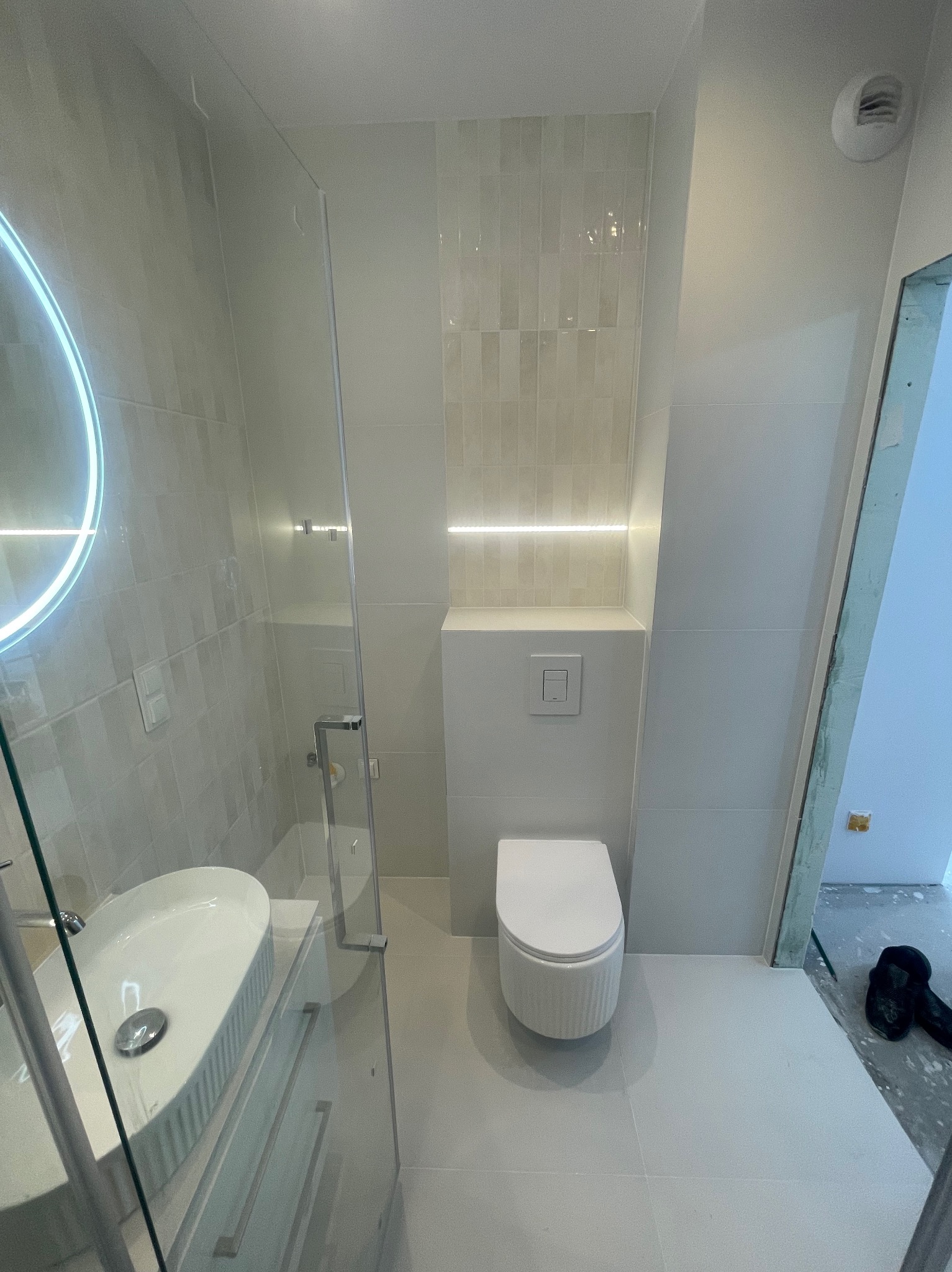 Nowoczesna łazienka w jasnych odcieniach szarości z okrągłym lustrem z podświetleniem LED, wiszącą toaletą i szklaną kabiną prysznicową.