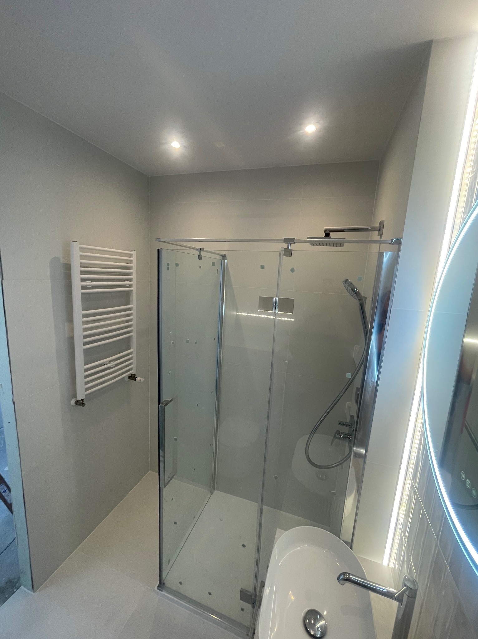 Nowoczesna łazienka z kabiną prysznicową ze szklanymi drzwiami, grzejnikiem drabinkowym i okrągłym lustrem z podświetleniem LED. Jasne płytki i minimalistyczny design.