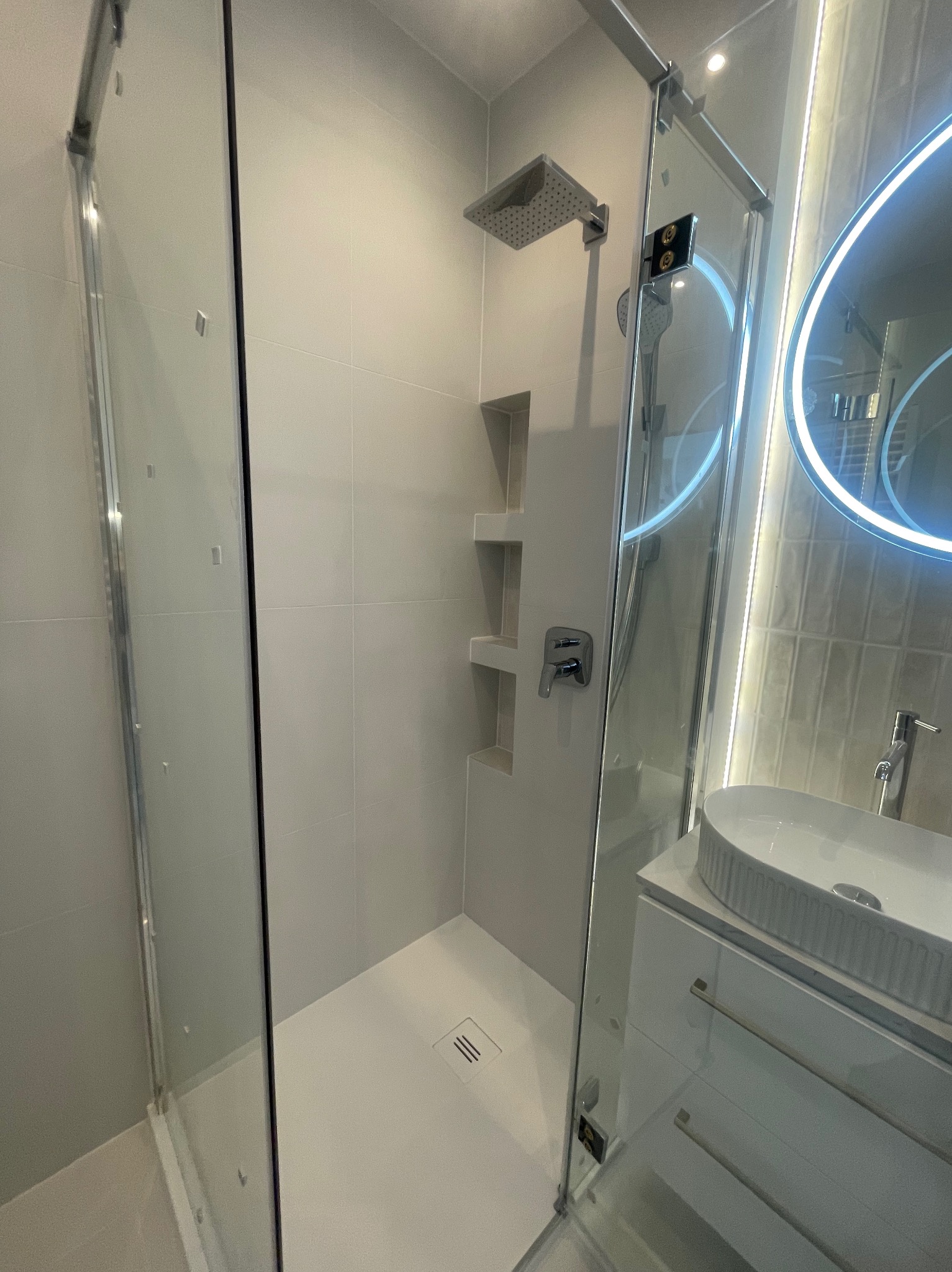 Nowoczesna kabina prysznicowa walk-in z wnękami na kosmetyki, szklane drzwi, biała armatura i okrągłe lustro z podświetleniem LED. Minimalistyczny design.