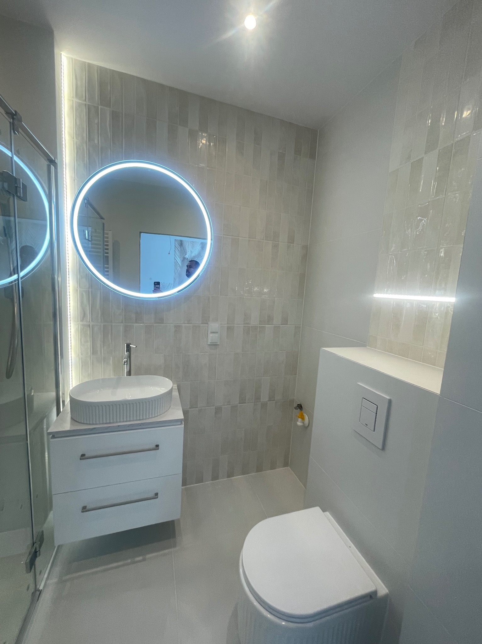 Nowoczesna łazienka w odcieniach szarości z okrągłym lustrem LED, umywalką nablatową i podwieszaną toaletą. Minimalistyczny design, płytki imitujące kamień, subtelne oświetlenie.