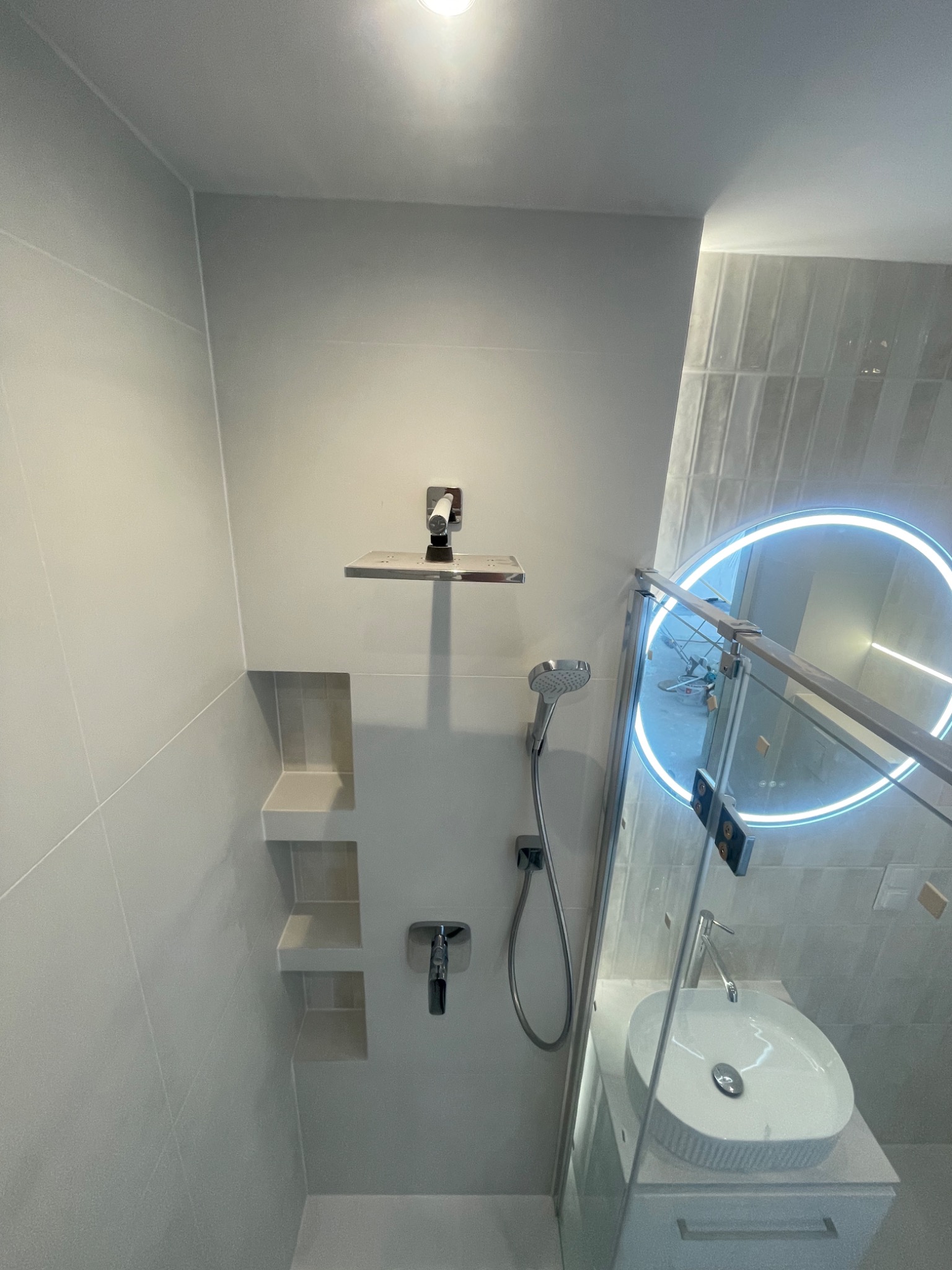 Nowoczesna łazienka po remoncie: szare płytki, wnęki w ścianie, deszczownica, umywalka z podświetlanym lustrem i szklany panel prysznicowy.