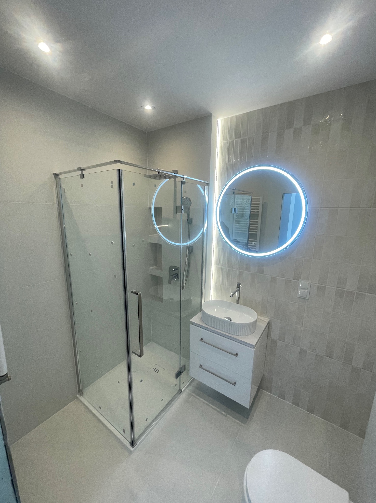 Nowoczesna łazienka w szarościach z kabiną prysznicową, okrągłym lustrem z podświetleniem LED i wiszącą szafką z umywalką. Minimalistyczny design.