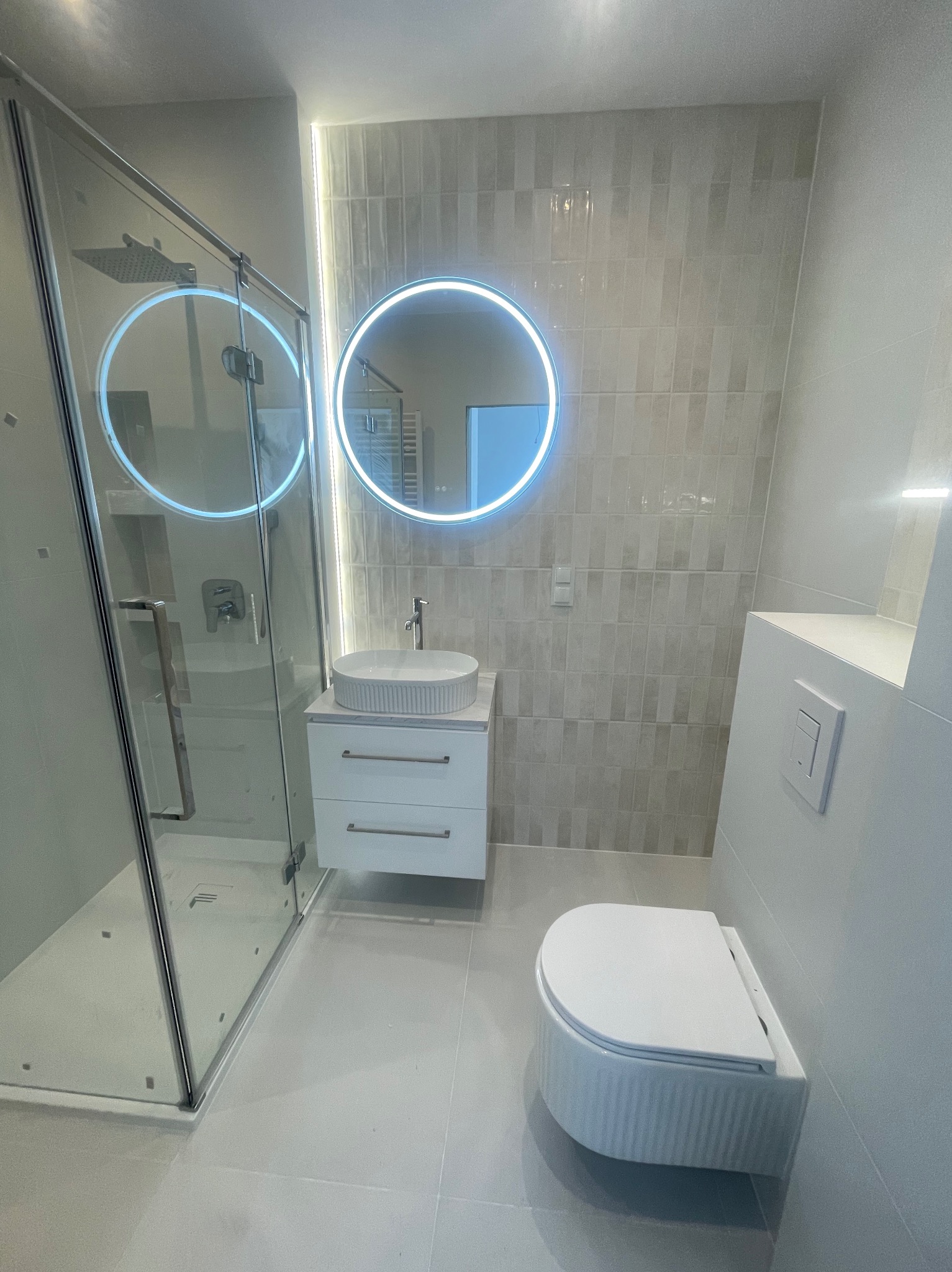 Nowoczesna łazienka w odcieniach szarości z podświetlanym lustrem, kabiną prysznicową i podwieszaną toaletą. Minimalistyczny design, płytki imitujące cegłę.