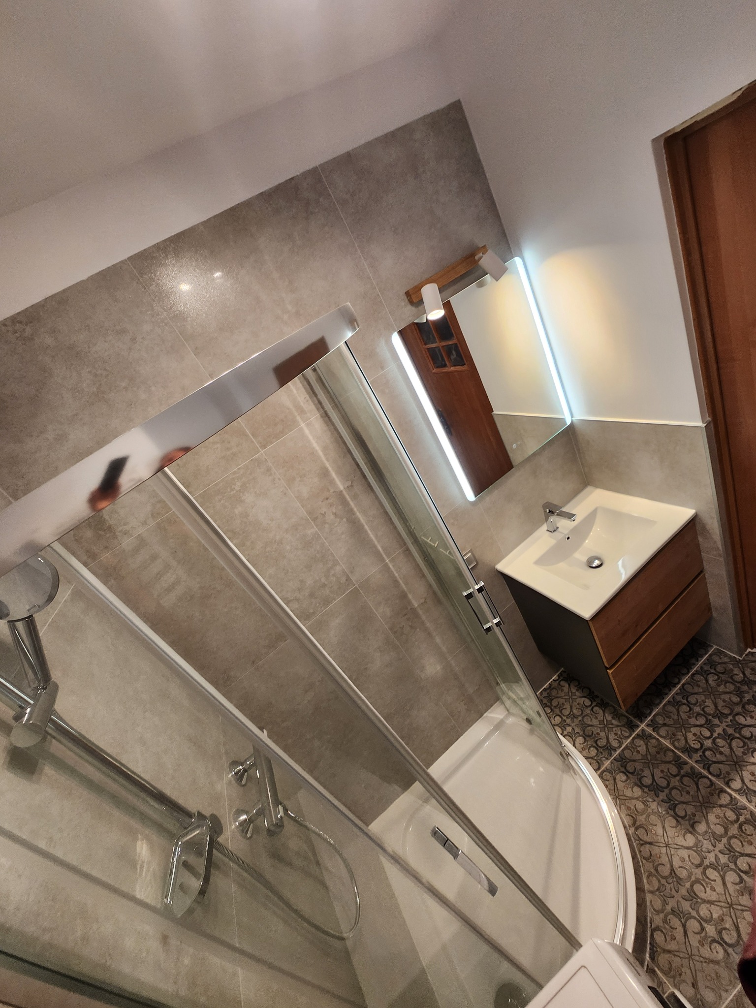 Nowoczesna łazienka po remoncie: kabina prysznicowa z chromowanymi elementami, umywalka z drewnianą szafką i podświetlane lustro. Płytki w odcieniach szarości i wzorzysta podłoga.