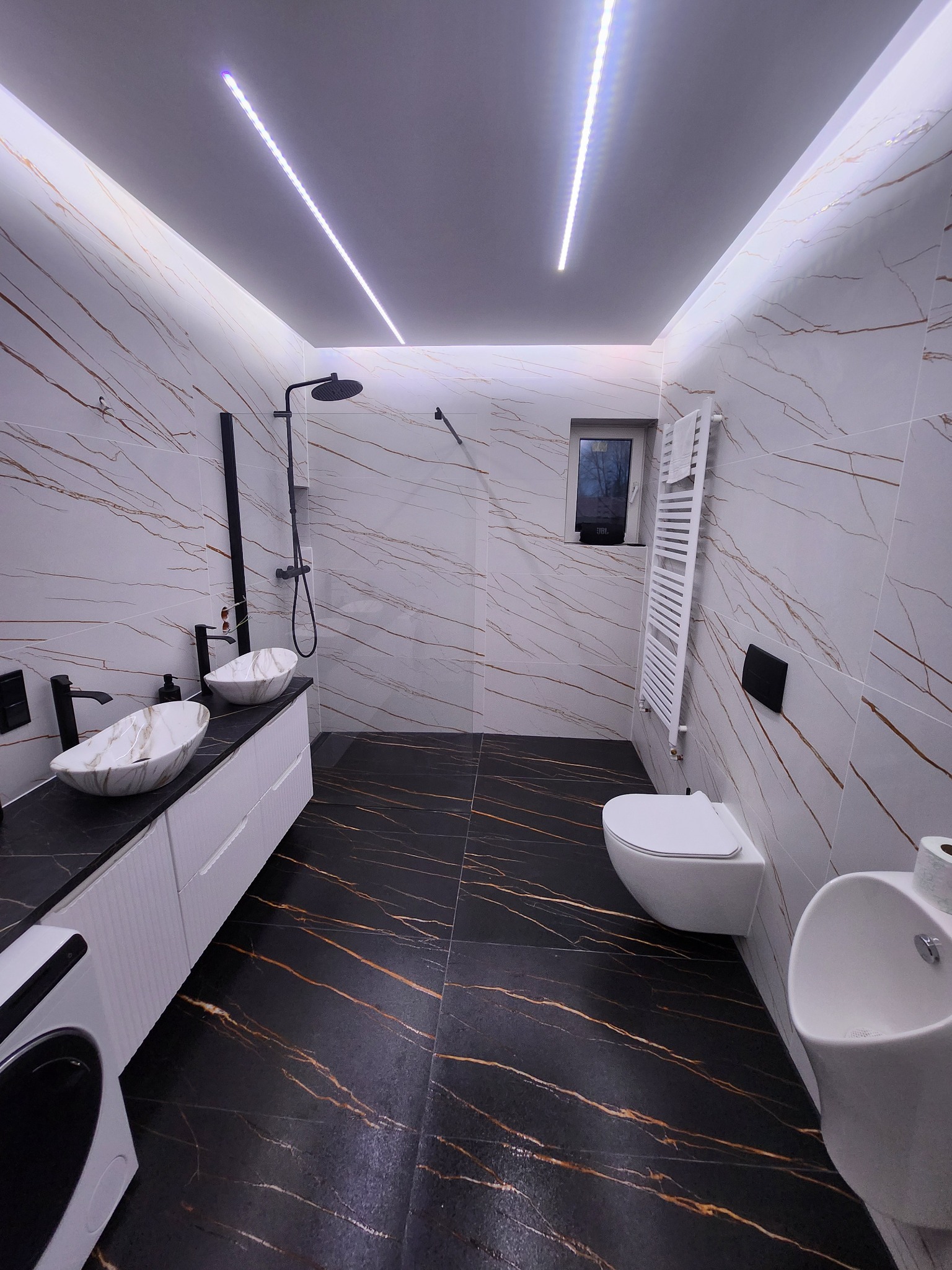 Nowoczesna łazienka z czarno-złotą podłogą i białymi marmurkowymi ścianami. Dwie umywalki na blacie, czarna armatura, oświetlenie LED i grzejnik drabinkowy.