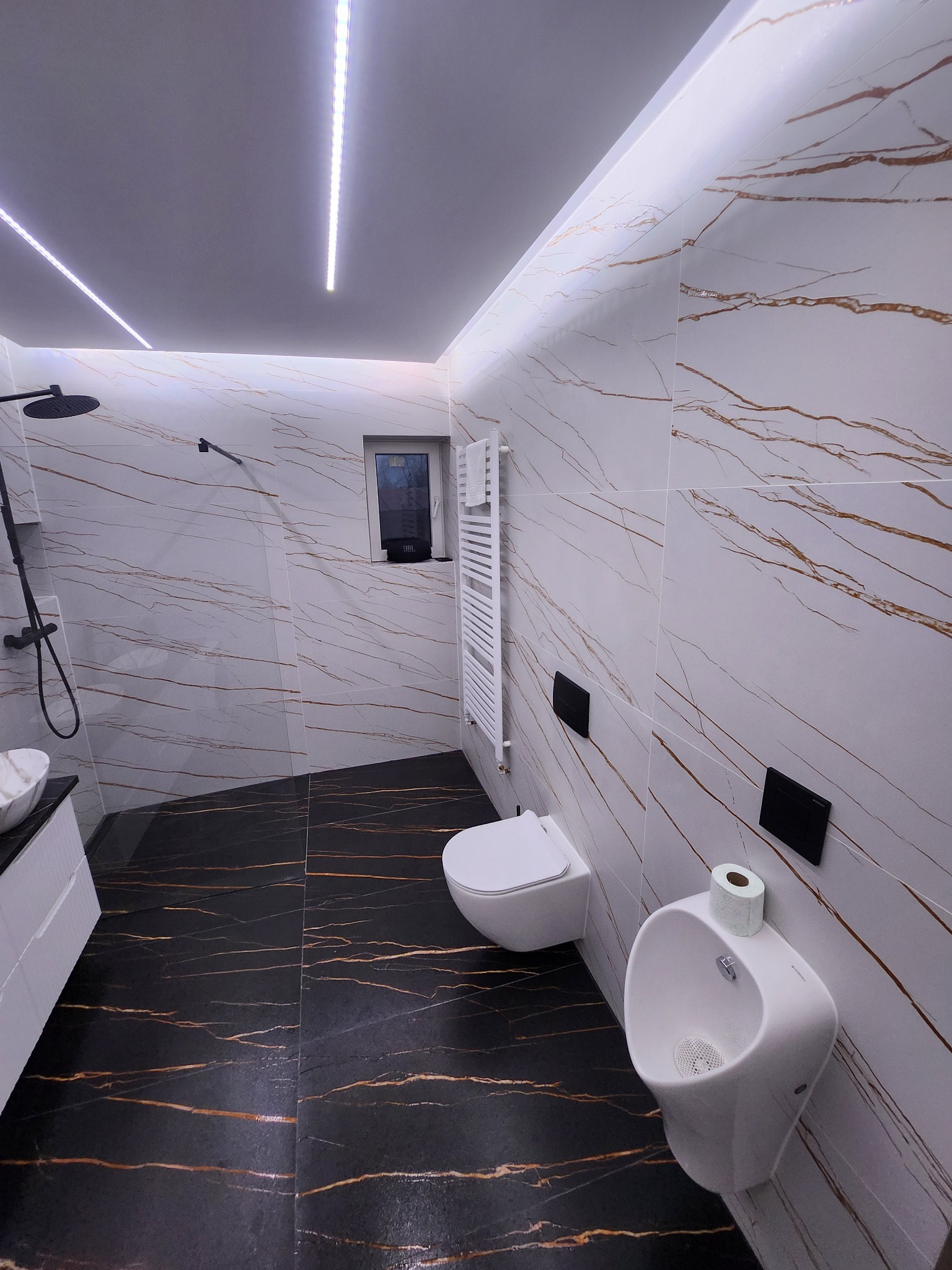 Nowoczesna łazienka z czarno-złotą podłogą i białymi płytkami imitującymi marmur. Widoczna umywalka, toaleta, pisuar i grzejnik drabinkowy. Oświetlenie LED na suficie.