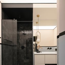 Latre Design - Nowoczesna łazienka z czarnymi płytkami w kabinie prysznicowej, złotą lampą wiszącą nad umywalką i beżowymi szafkami.