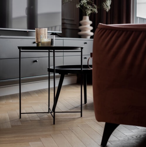 Minimalistyczny salon: czarny stolik kawowy z dekoracjami (świece, wazon) na tle szarej komody i parkietu w jodełkę, z fragmentem ceglastego fotela w kadrze.