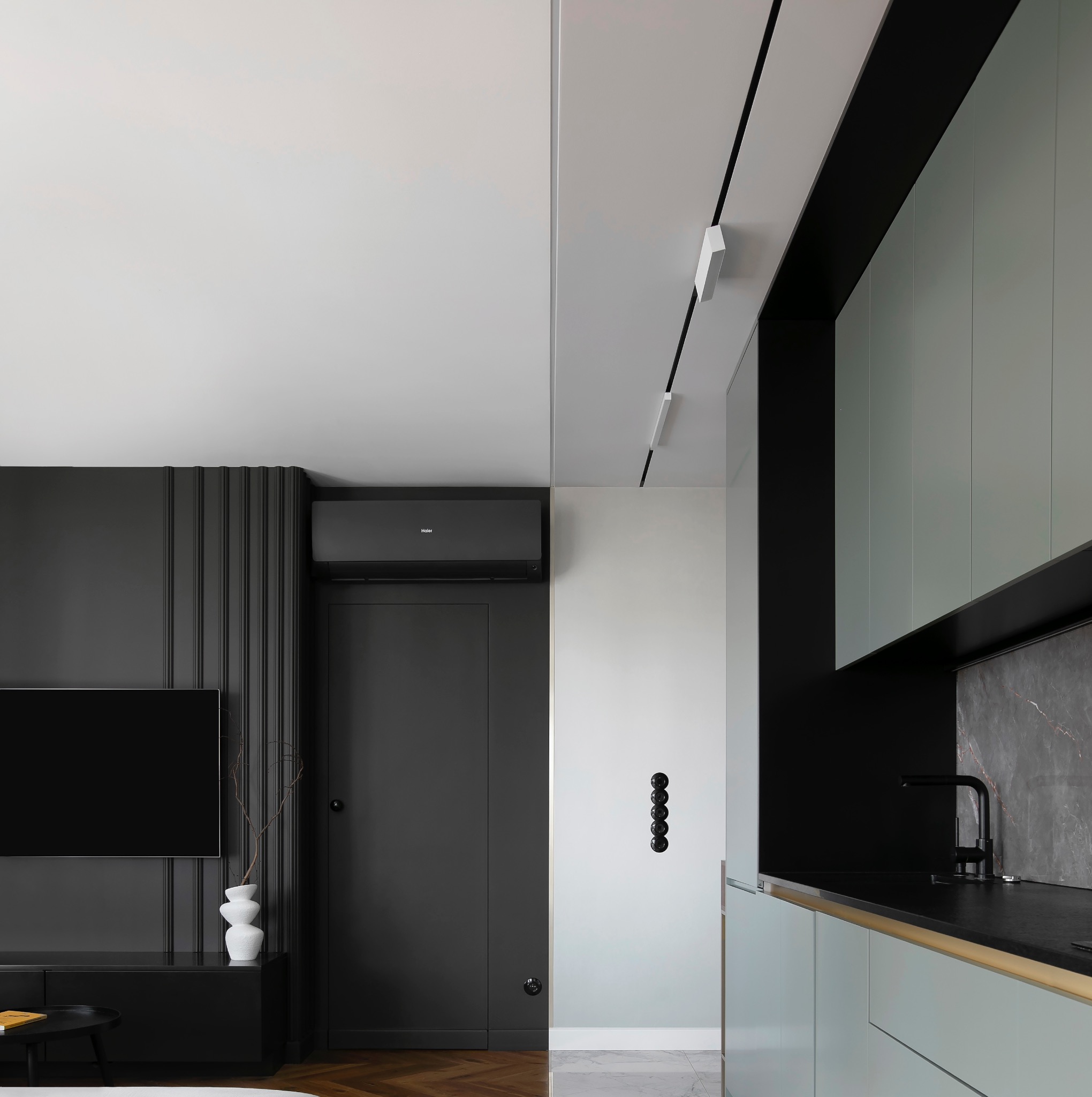 Nowoczesne wnętrze mieszkania w stonowanej kolorystyce: czarna ściana z telewizorem i dekoracyjnym wazonem, minimalistyczna kuchnia z szarymi szafkami i czarnym blatem, parkiet w jodełkę...