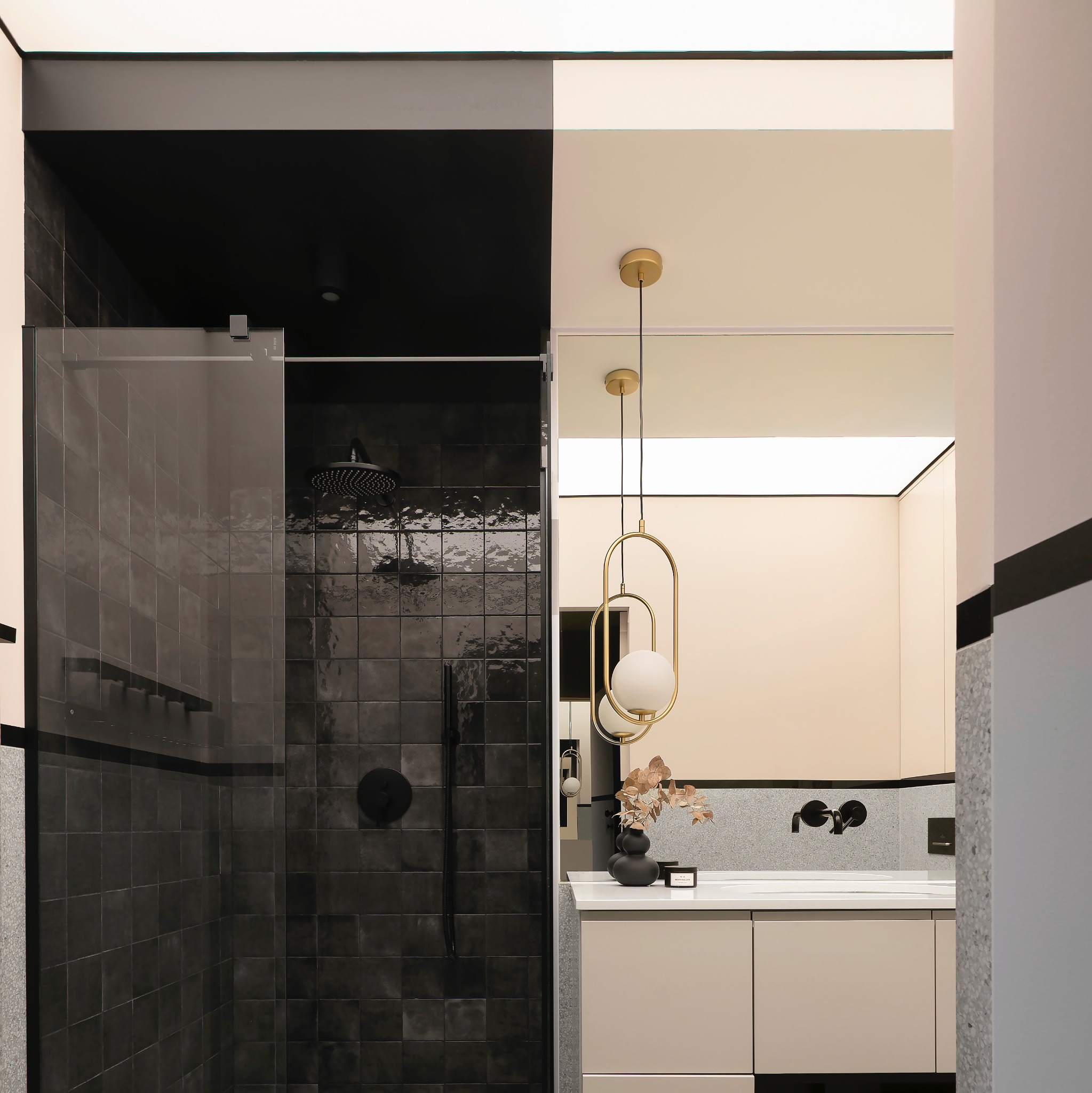 Nowoczesna łazienka z czarnymi płytkami w kabinie prysznicowej, złotą lampą wiszącą nad umywalką i beżowymi szafkami.