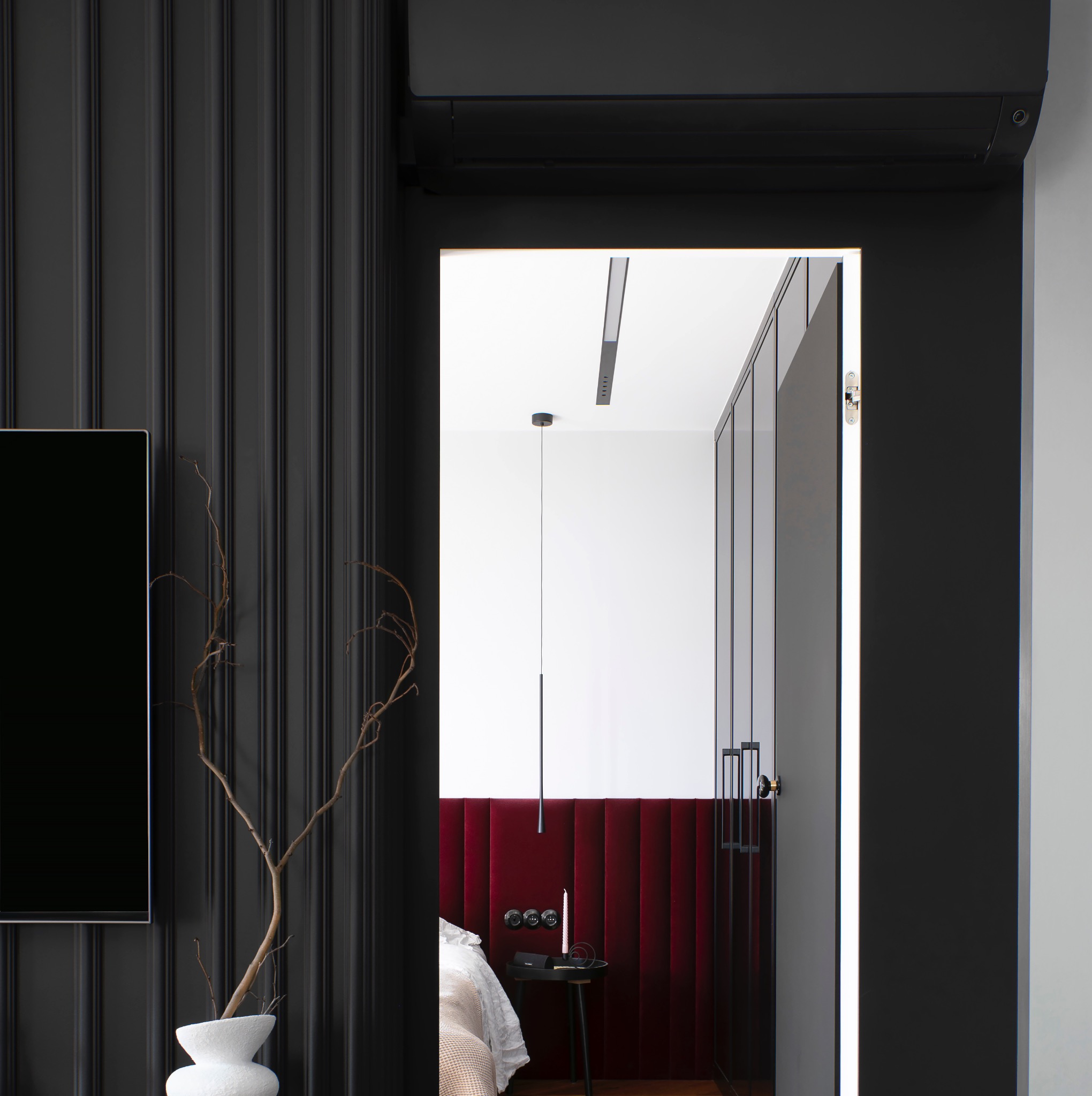 Nowoczesna sypialnia z ciemną ścianą z listwami, telewizorem i dekoracyjnym suchym bukietem w białym wazonie, widok zza czarnych drzwi na łóżko z bordowym zagłówkiem i minimalistyczną lampą wiszącą.