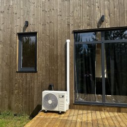 Thermo-Flow - Biały klimatyzator zewnętrzny zamontowany na drewnianym tarasie przy ścianie domu z drewnianą elewacją i czarnymi oknami. Widoczna instalacja.