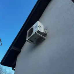 Biały klimatyzator Fujitsu zamontowany na szarej elewacji domu pod ciemnym okapem dachu, na tle błękitnego nieba. Widok z dołu.