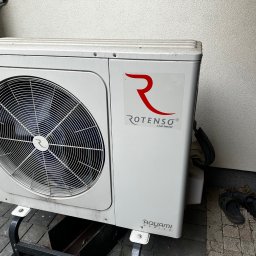 Thermo-Flow - Biała jednostka zewnętrzna klimatyzacji Rotenso Aquami Series na metalowej podstawie, ustawiona na kostce brukowej przy ścianie. Obok leżą klapki.