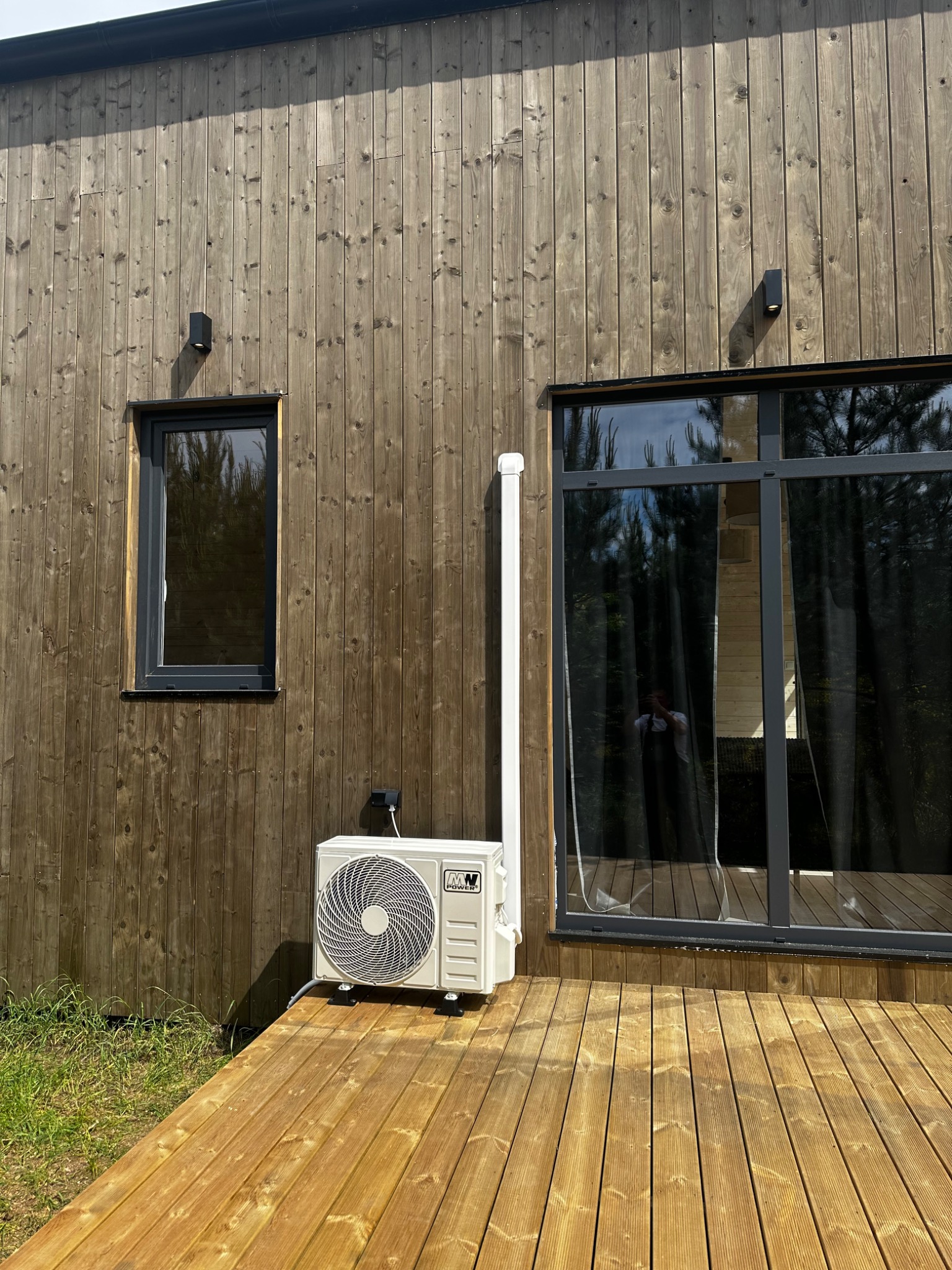 Biały klimatyzator zewnętrzny zamontowany na drewnianym tarasie przy ścianie domu z drewnianą elewacją i czarnymi oknami. Widoczna instalacja.