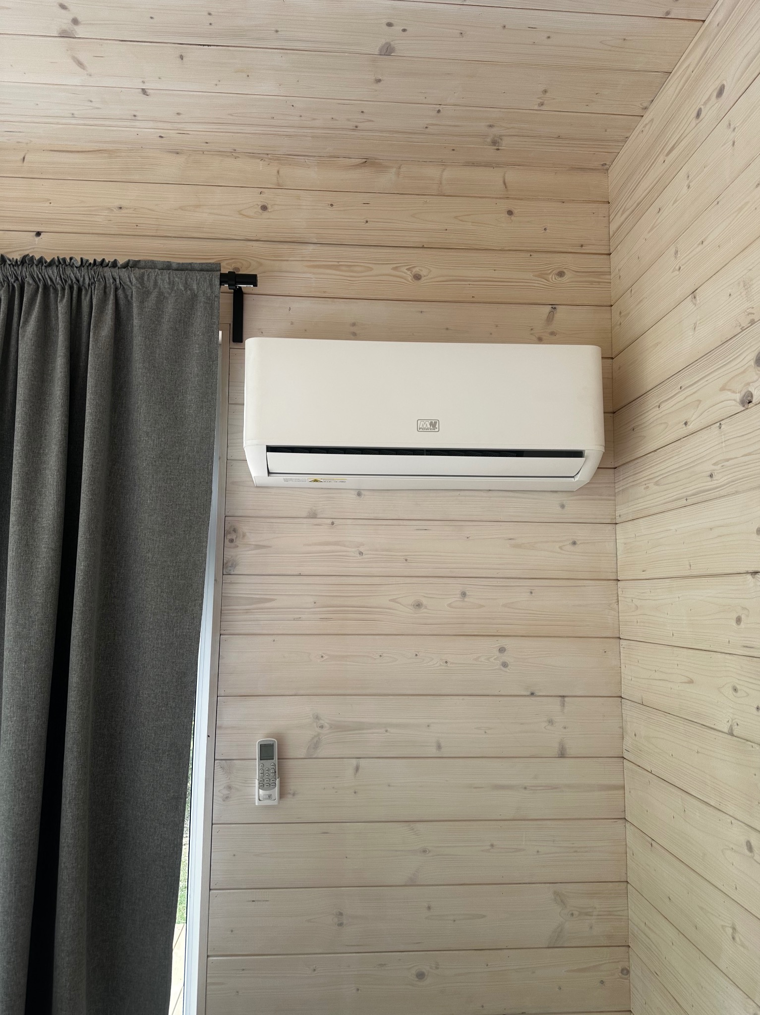 Biały klimatyzator ścienny zamontowany na drewnianej ścianie, obok okna z szarą zasłoną i pilotem. Wnętrze wykończone boazerią w jasnym odcieniu.