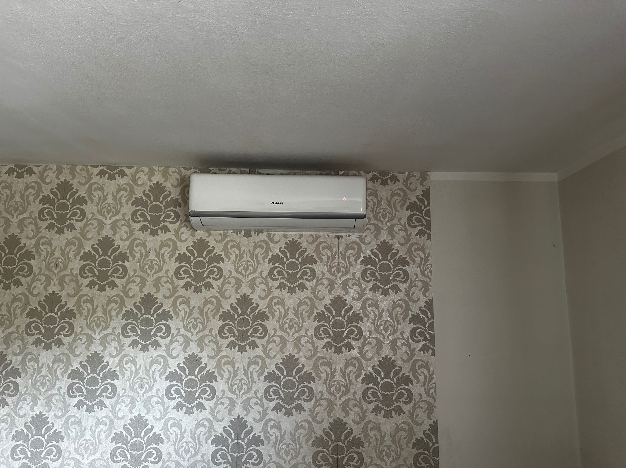 Biała klimatyzacja marki Gree zamontowana na ścianie z tapetą w kwiaty, widok od dołu. Świecąca dioda sygnalizuje pracę urządzenia.
