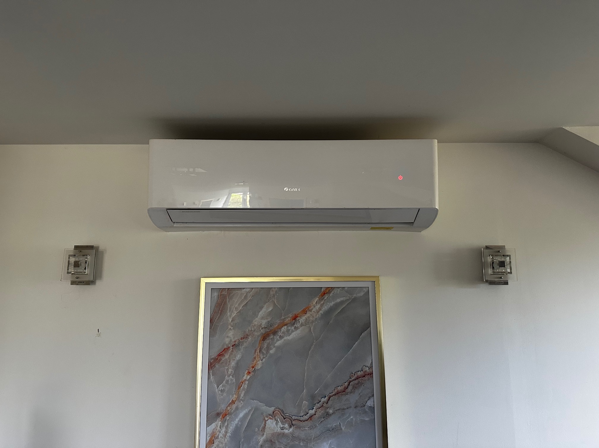 Biała klimatyzacja ścienna marki Gree zamontowana w pokoju z obrazem w złotej ramie i dwoma kinkietami. Widoczny fragment sufitu.