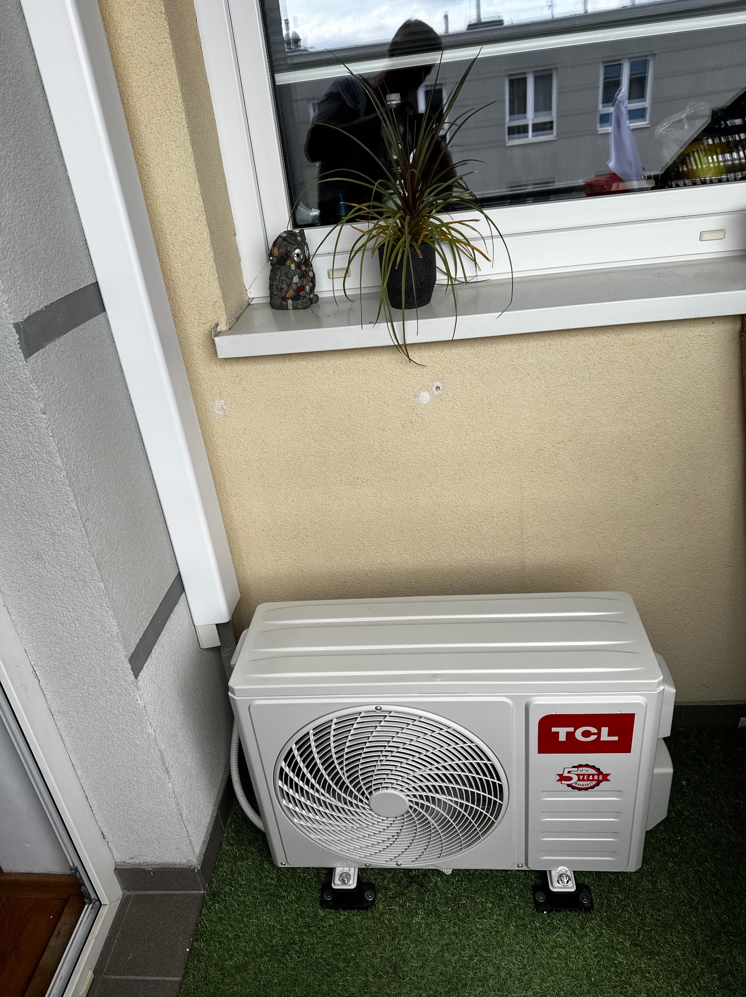 Biała jednostka zewnętrzna klimatyzacji TCL zamontowana na balkonie z sztuczną trawą, widoczna z boku, z oknem w tle, obok dekoracyjna figurka sowy.