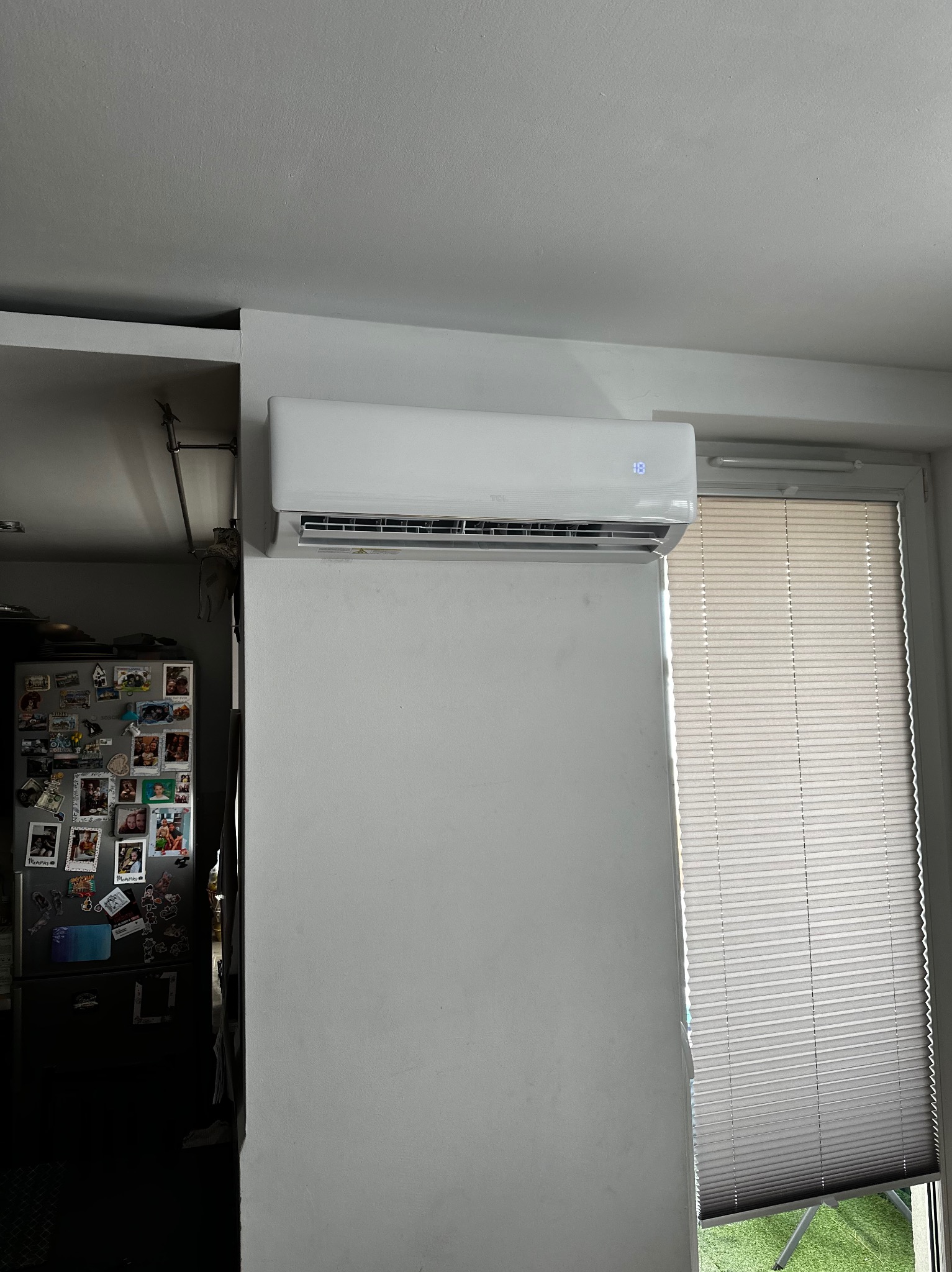 Biały klimatyzator ścienny z wyświetlaczem temperatury 18 stopni, zamontowany w mieszkaniu obok okna z żaluzjami i lodówki z magnesami.