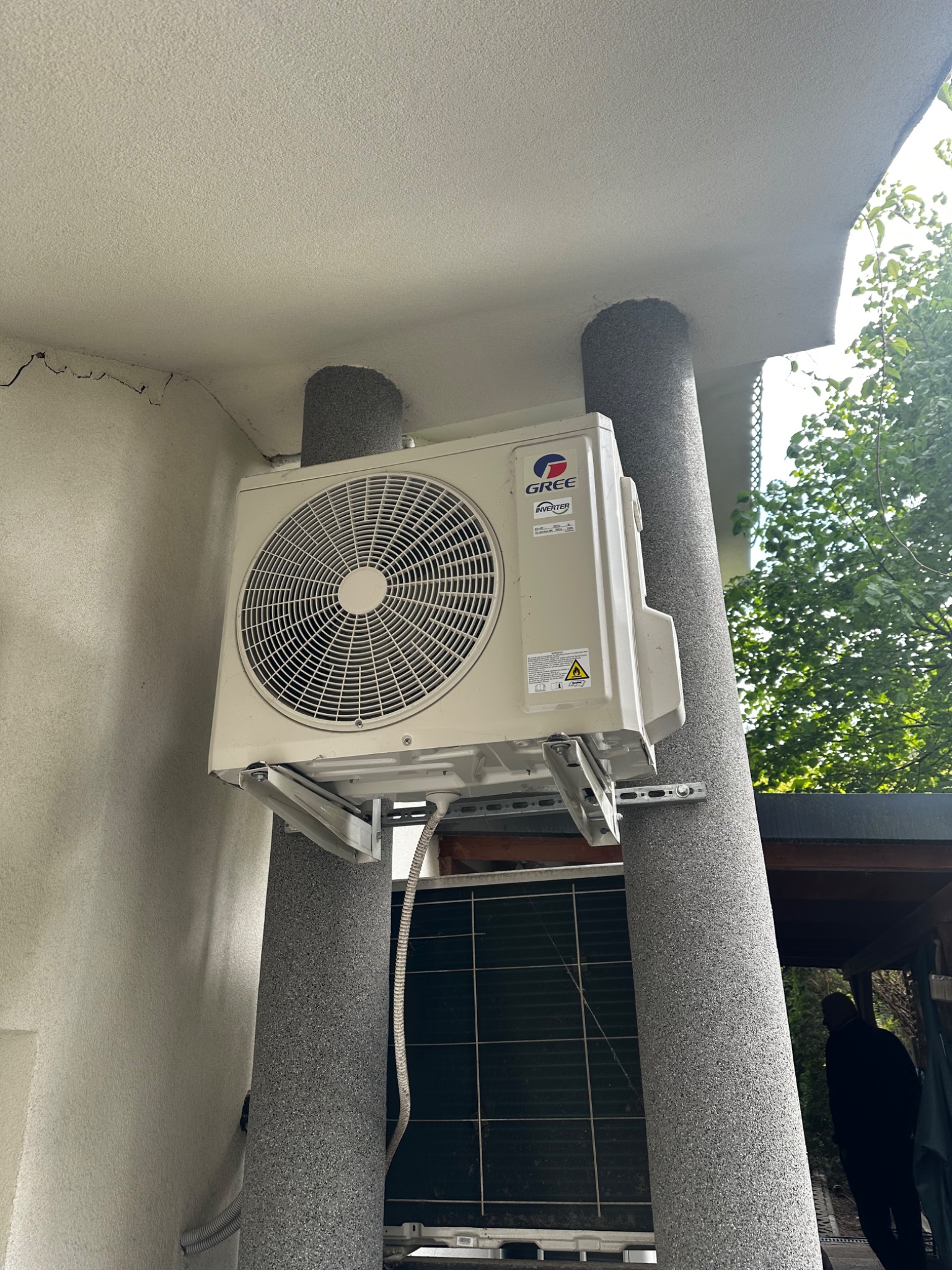 Zamontowana jednostka zewnętrzna klimatyzacji Gree na szarej elewacji, widoczna dolna część drugiej jednostki. Ujęcie z dołu, fragment drzewa w tle.