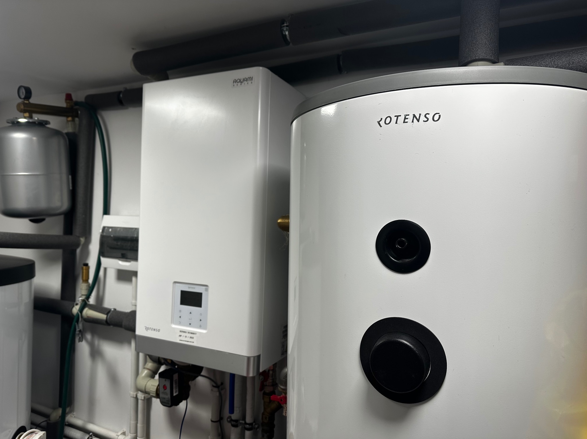 Biała pompa ciepła Aquami i zasobnik Rotenso w kotłowni, widoczne rury instalacyjne i elementy armatury. Całość w jasnej, minimalistycznej aranżacji.
