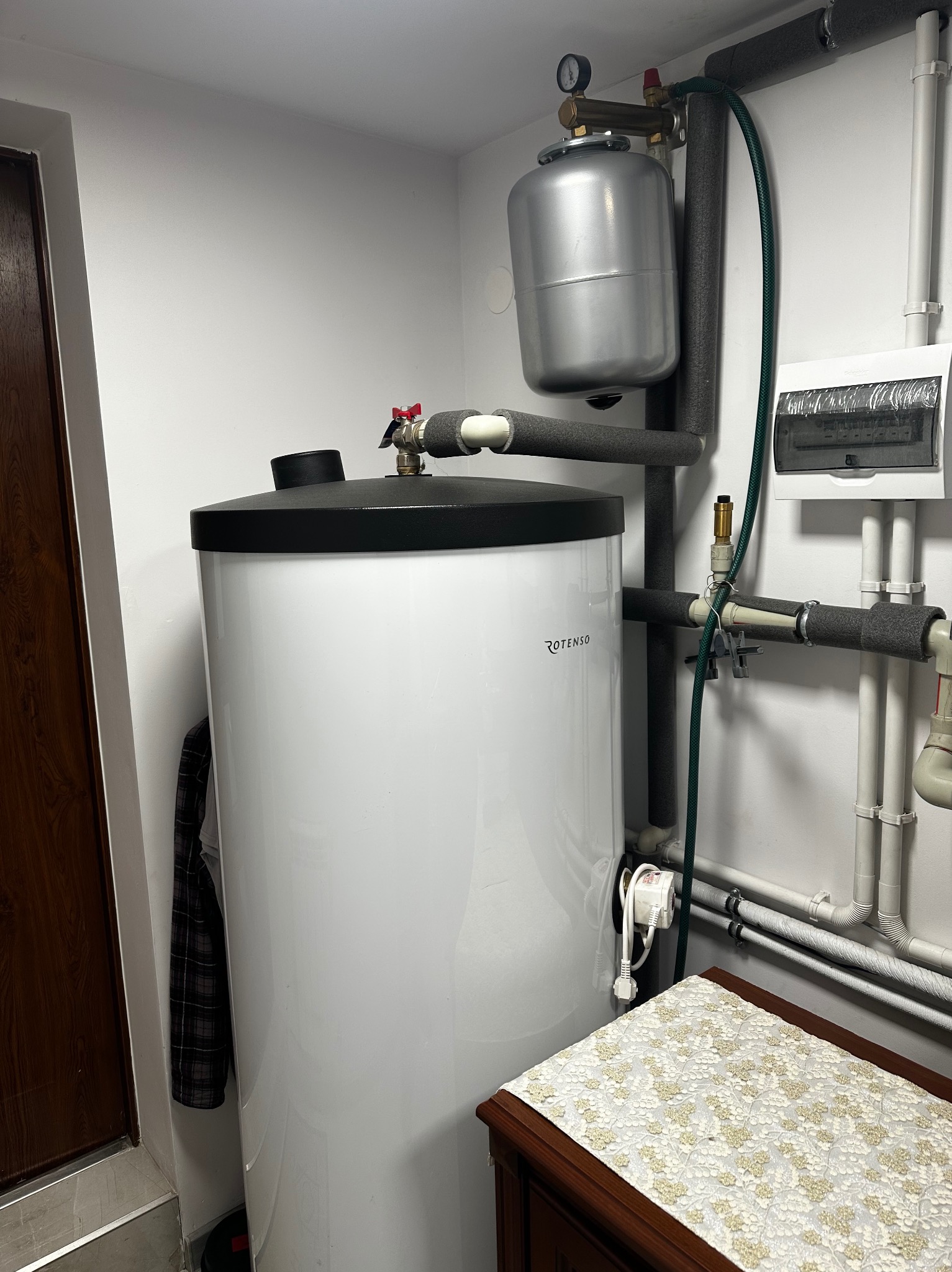 Biały, cylindryczny zbiornik Rotenso z czarną górą, część instalacji grzewczej w pomieszczeniu z rurami, zaworami i rozdzielnicą. Widoczny fragment szafki z ozdobnym obrusem.