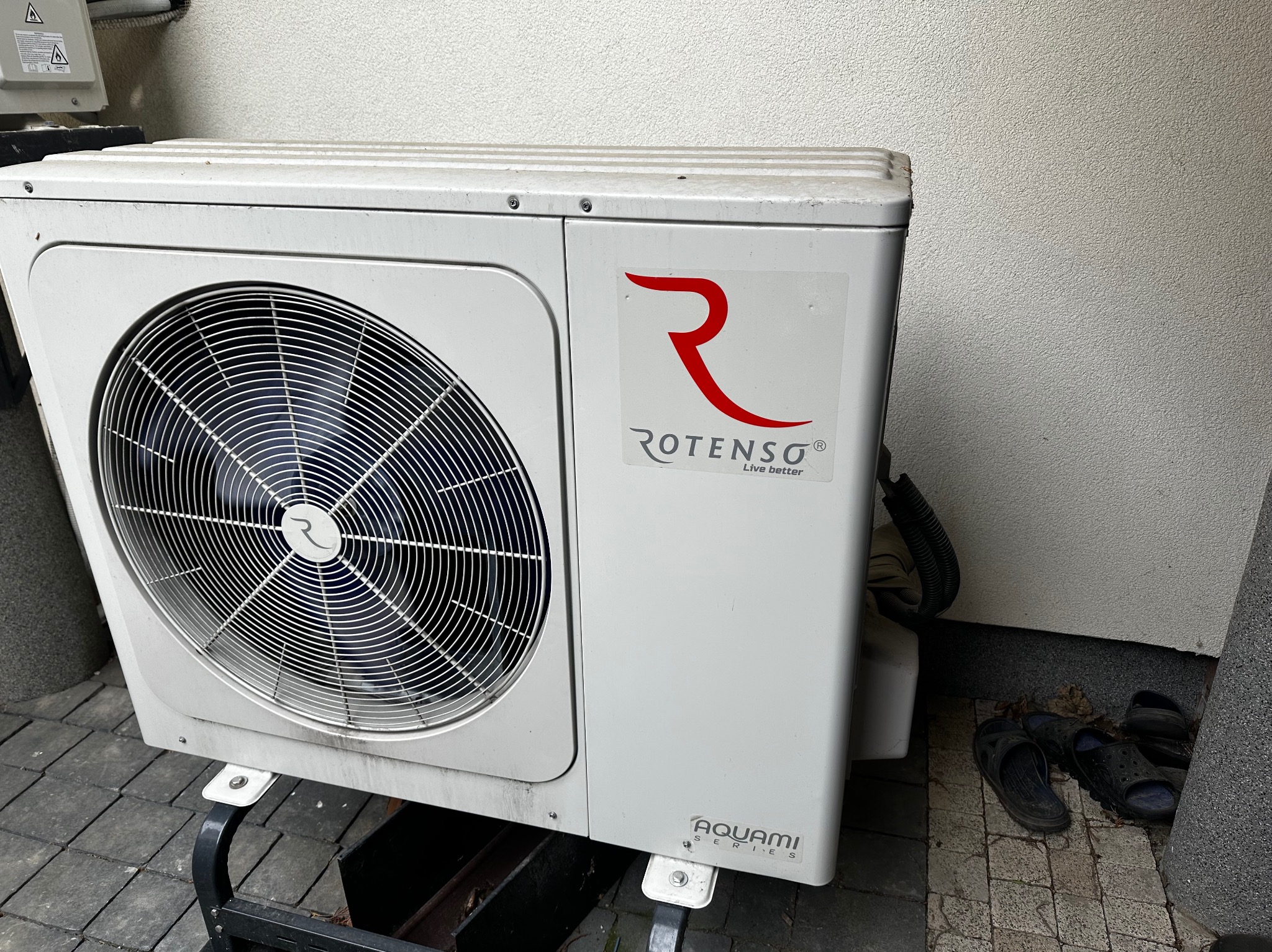 Biała jednostka zewnętrzna klimatyzacji Rotenso Aquami Series na metalowej podstawie, ustawiona na kostce brukowej przy ścianie. Obok leżą klapki.