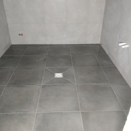 Mycie maszynowe - Wykończona szara łazienka z kwadratowymi płytkami i odpływem liniowym w podłodze, widoczne przyłącza wodne na ścianie.