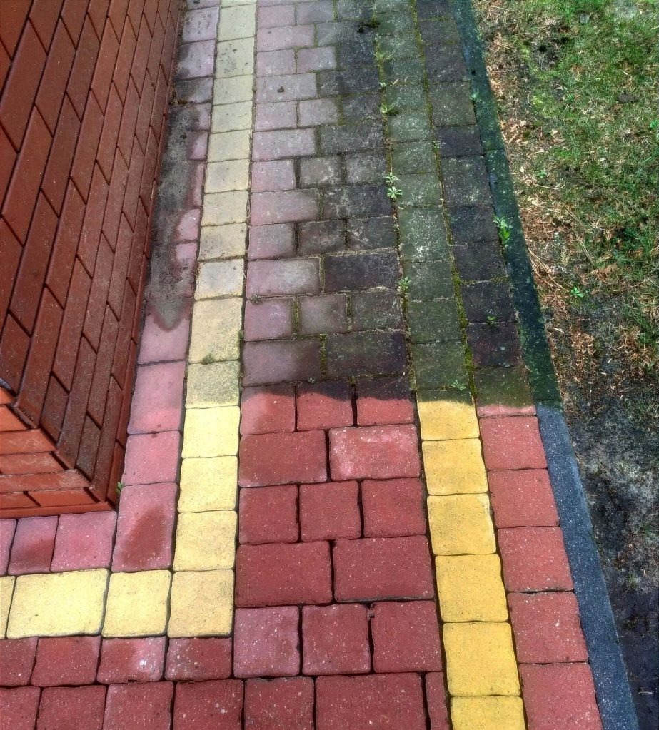 Częściowo wyczyszczona, kolorowa kostka brukowa przy ścianie budynku, widoczne różnice w czystości kostki.