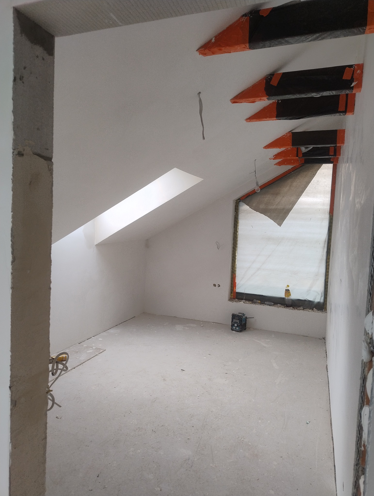 Pokój na poddaszu w trakcie prac wykończeniowych: surowe ściany, zakryte okno, widoczne kable, poziomica laserowa na podłodze, zabezpieczone elementy konstrukcyjne na suficie.