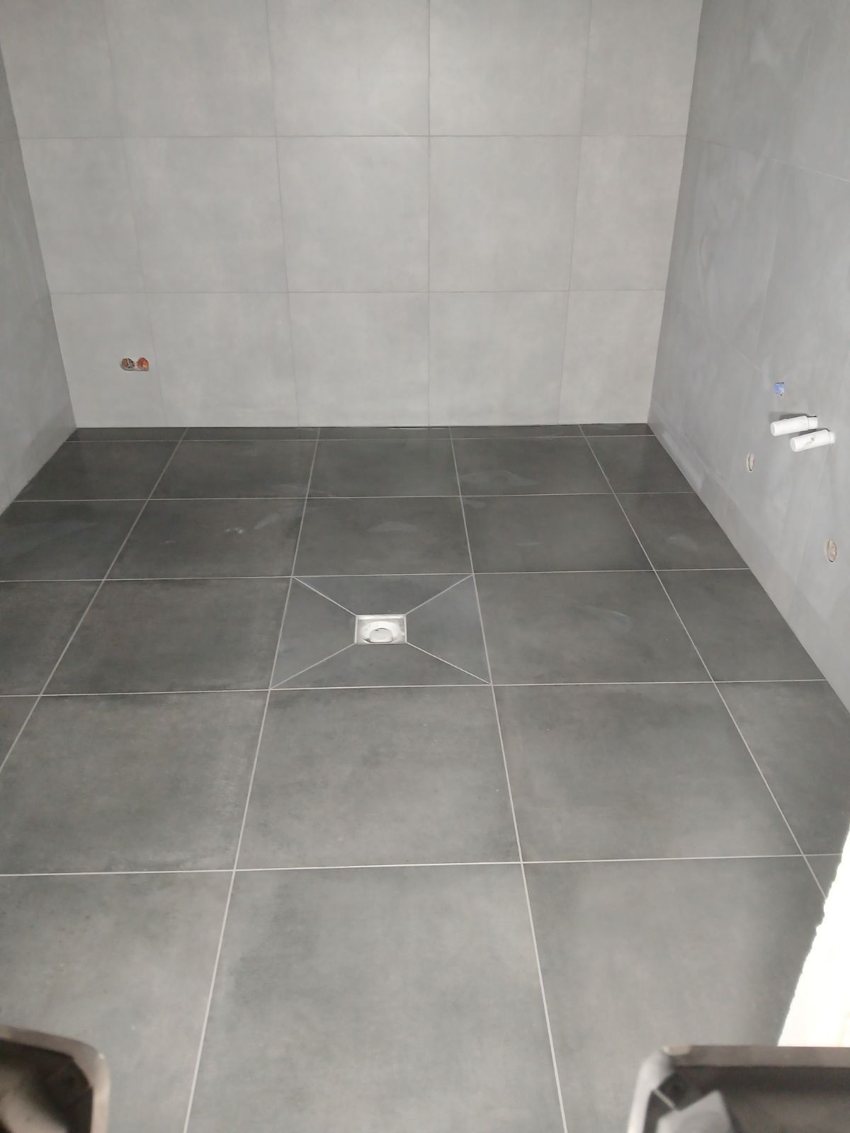 Wykończona szara łazienka z kwadratowymi płytkami i odpływem liniowym w podłodze, widoczne przyłącza wodne na ścianie.